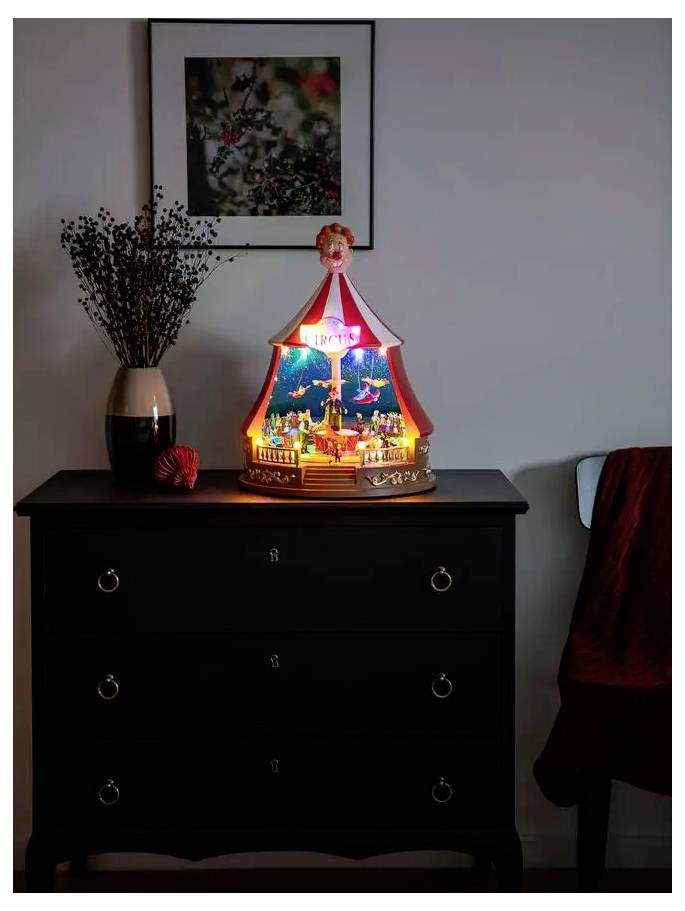 A lit miniature carousel holiday decoration sits on a dark dresser in a dim room, with a vase of branches and framed wall art nearby.