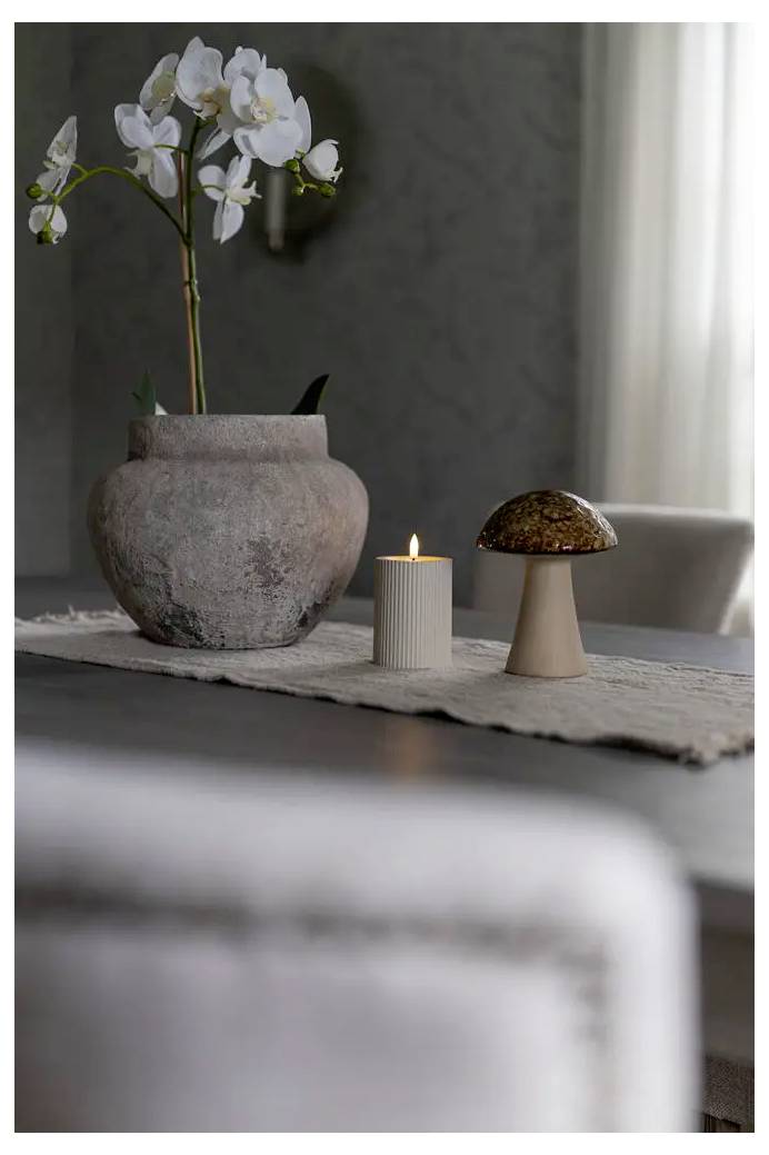 White orchid in a large ceramic pot beside a lit candle and small mushroom sculpture on a table runner in a softly lit room.