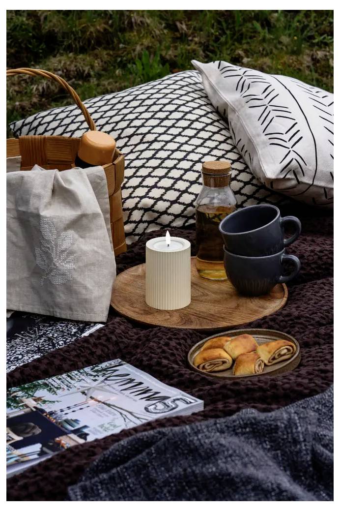 Picnic setup on a blanket with a basket, pillows, mugs, drink bottle, tea light, pastries, and a magazine outdoors.