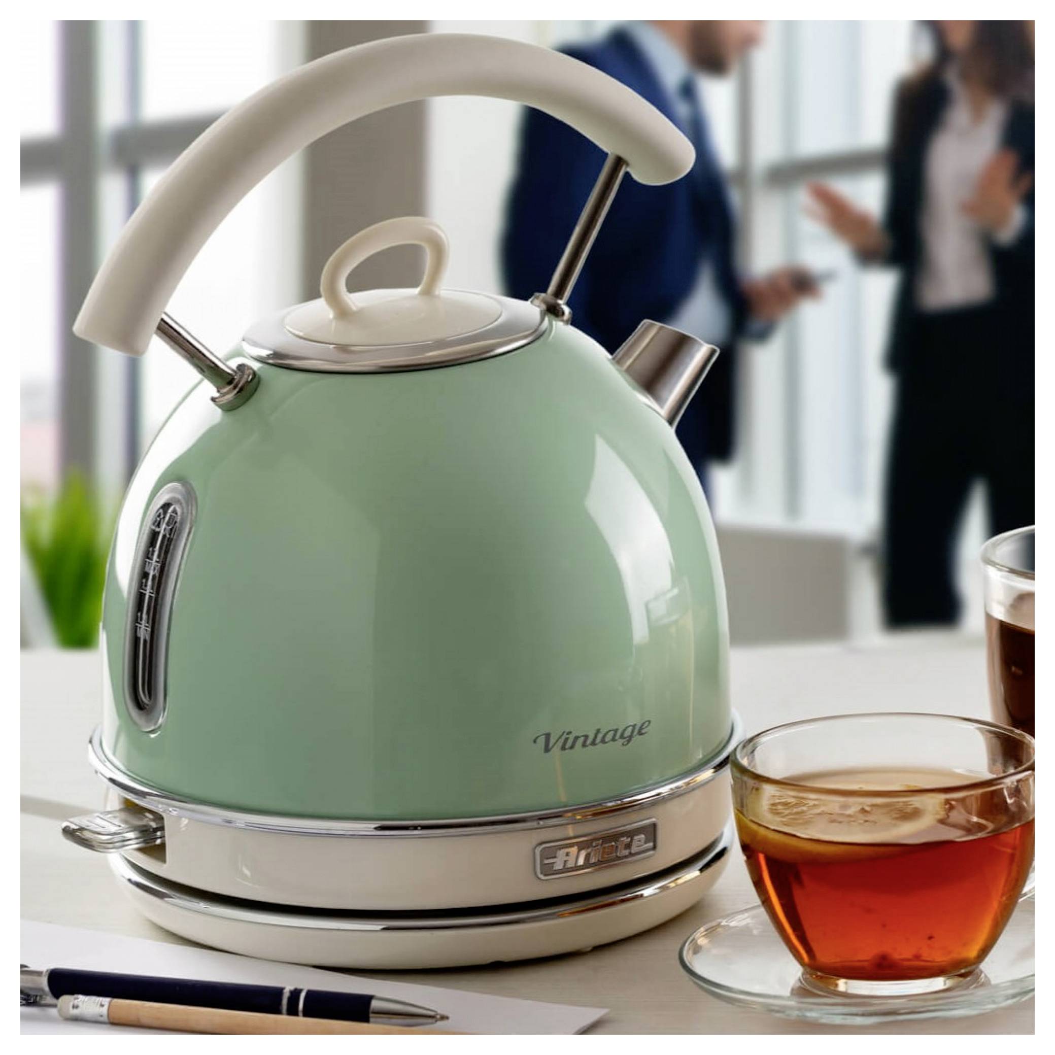 Mint-green vintage electric kettle on a table beside a cup of tea and a pen, with blurred people in the background.