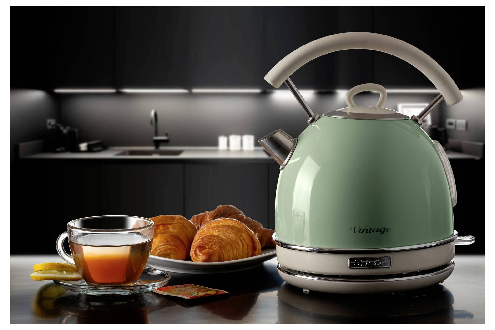 A mint-green electric kettle on a kitchen counter beside a glass of tea, croissants, and lemon slices in a modern dark kitchen.
