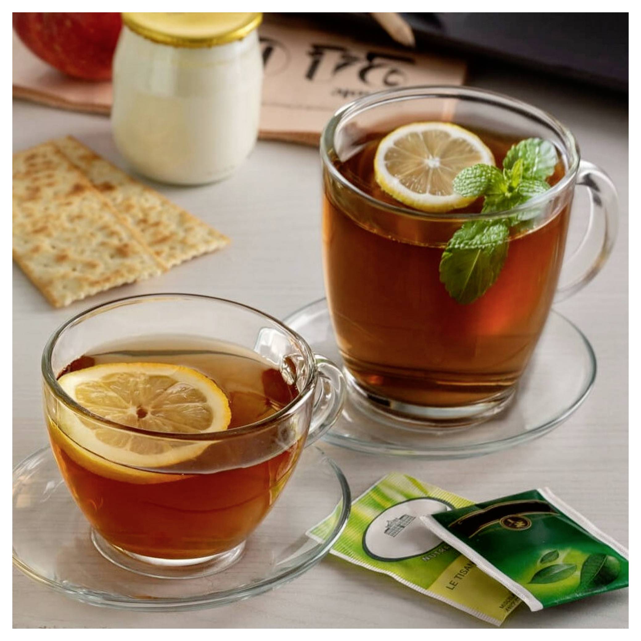 Two glass cups of hot tea with lemon slices, one garnished with mint, beside crackers and tea packets on a table.