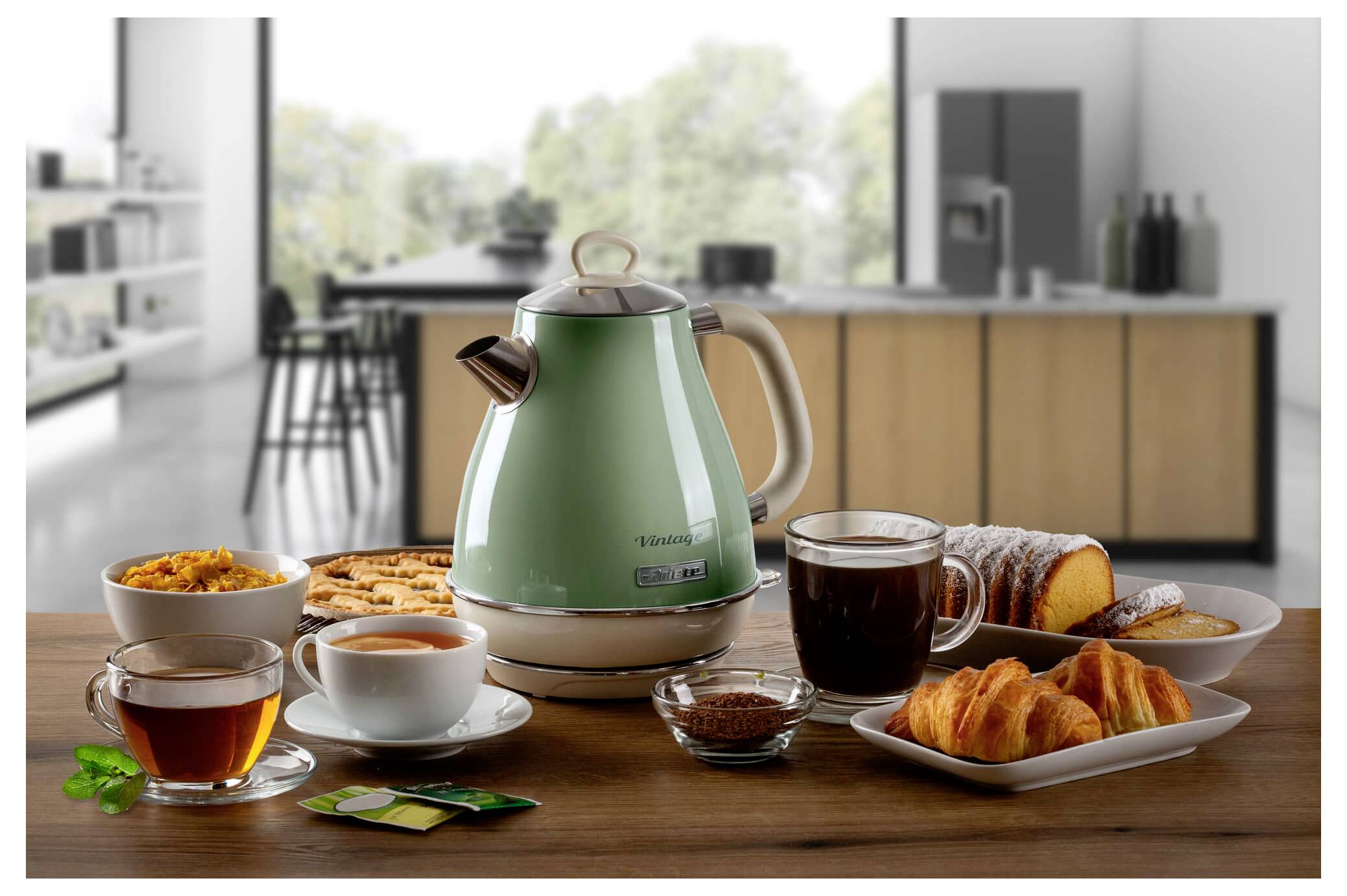 Breakfast spread on a wooden table with a green electric kettle, tea, coffee, pastries, and jam in a modern kitchen<br>