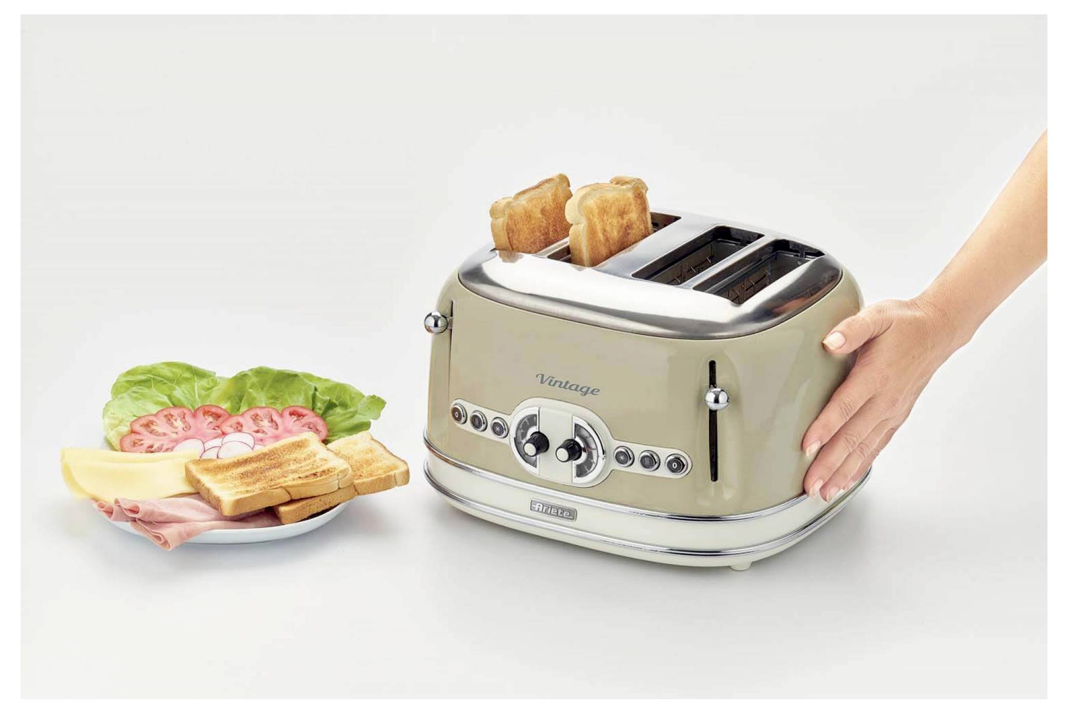 A hand places toast in a vintage four-slice toaster beside a plate of bread, deli meat, lettuce, and tomato on a white background.<br>