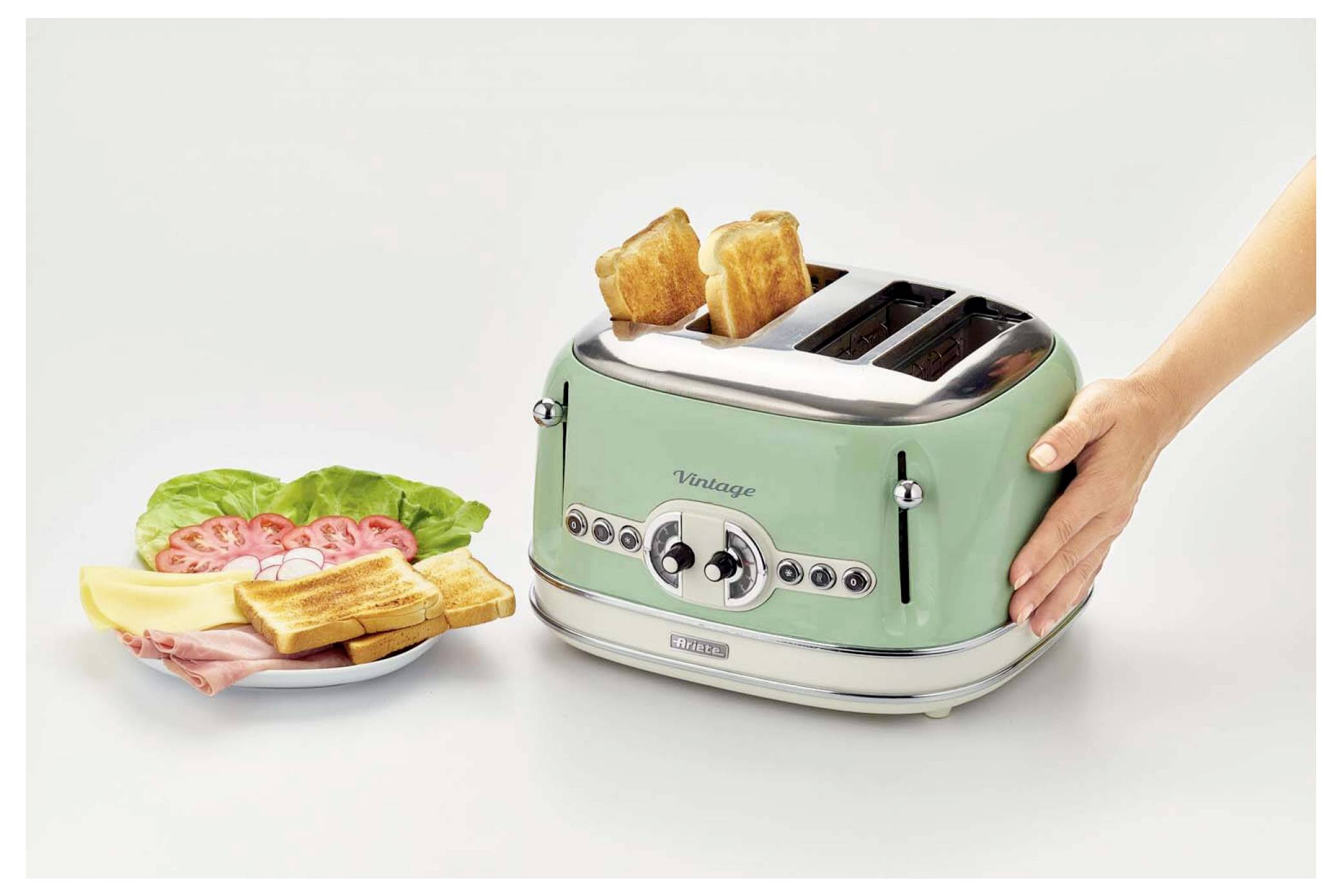 Hand placing toast in a mint-green vintage four-slot toaster beside a plate of breakfast toast with ham, lettuce, and tomato.