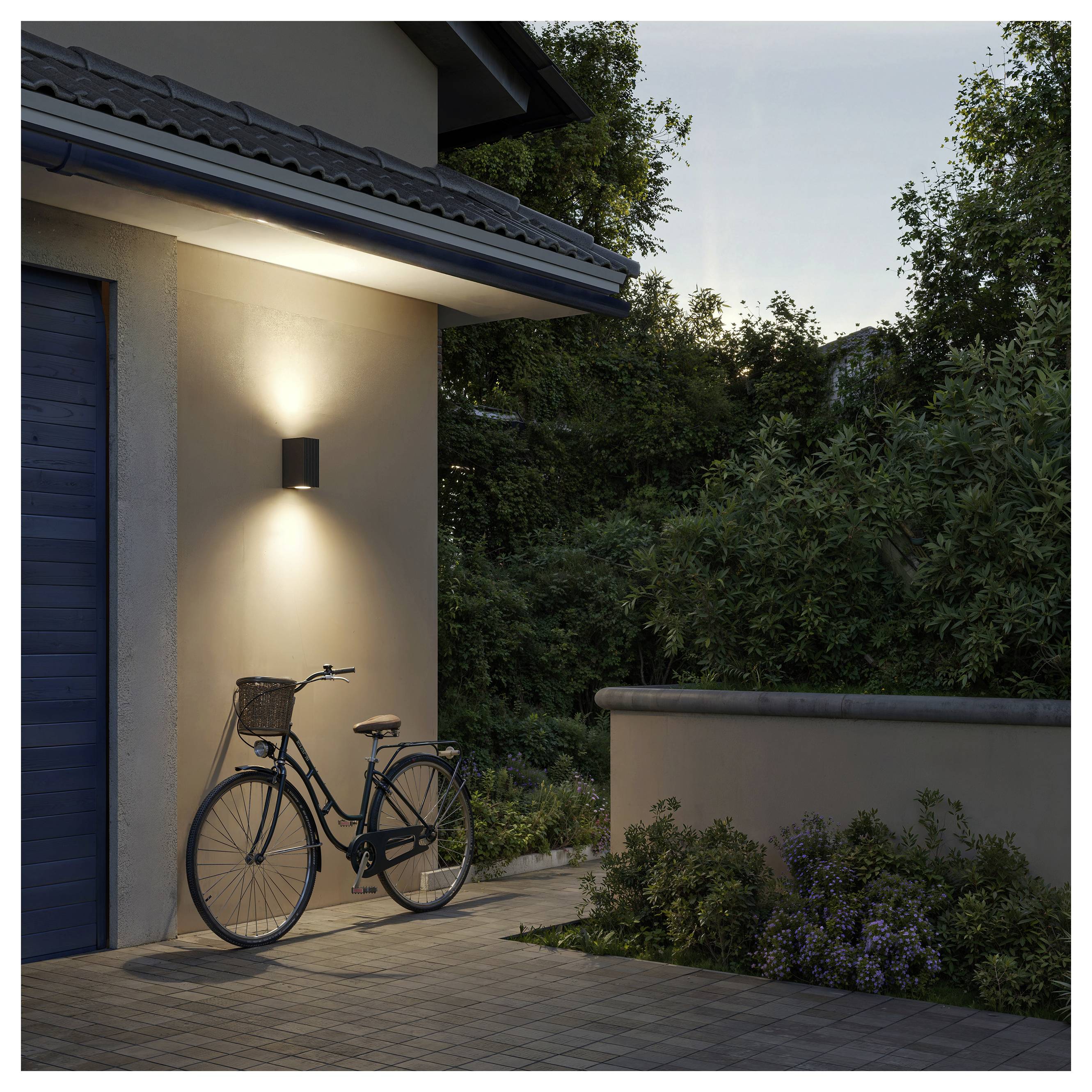 A bicycle leans against a house wall under a lit outdoor sconce at dusk, beside a patio and garden wall.