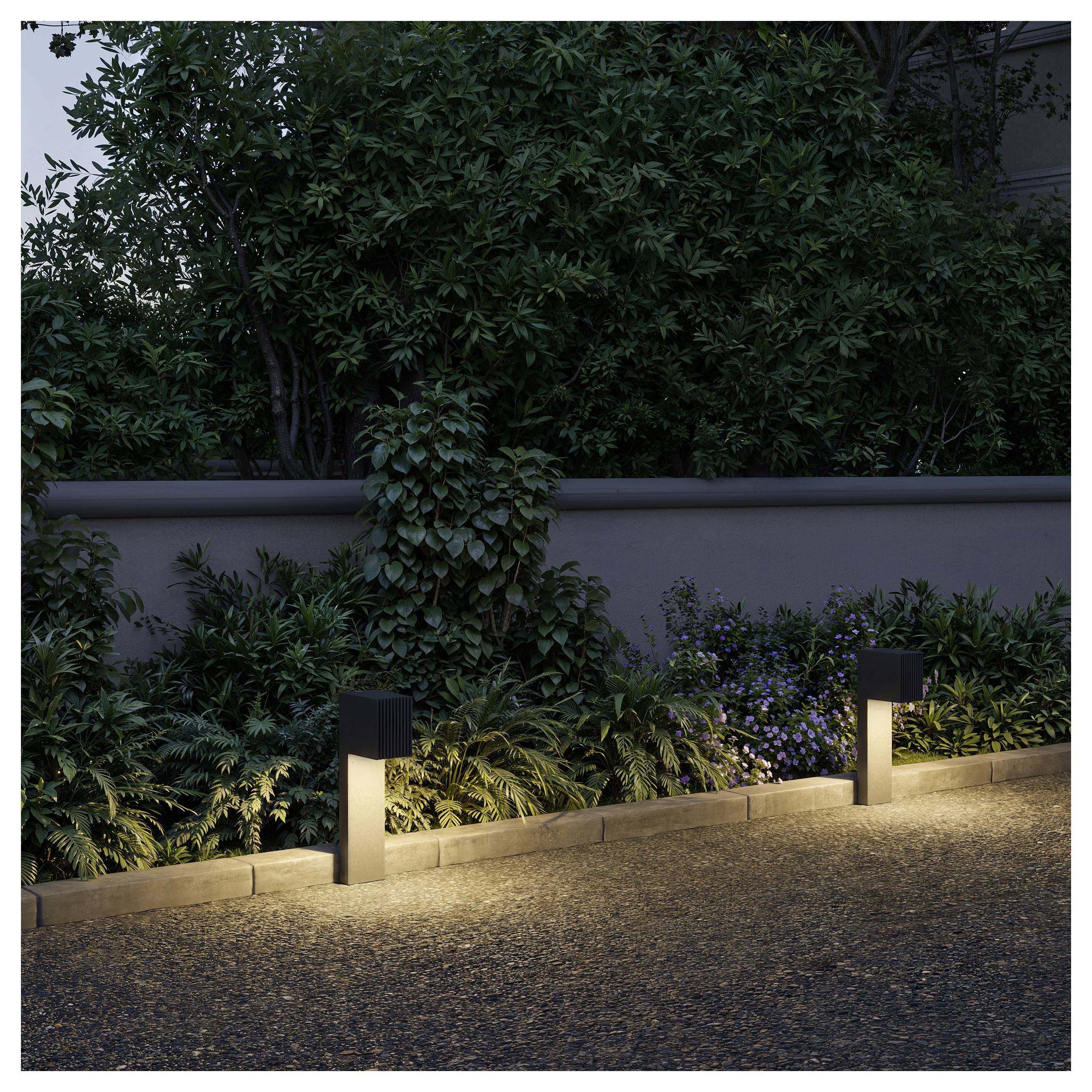 Two small black path lights illuminate a garden border with dense shrubs and a stone wall at dusk.