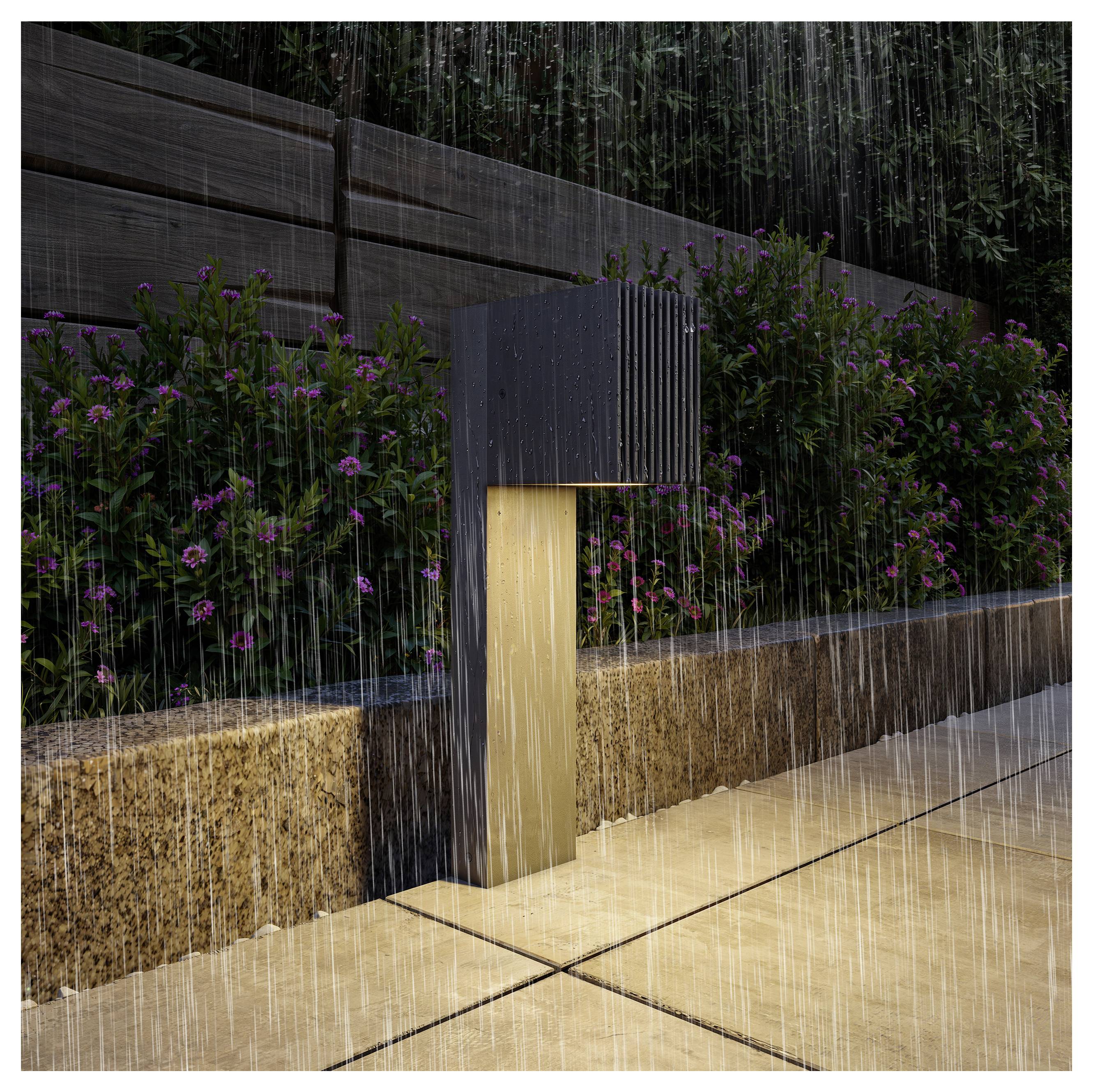 Modern outdoor light post glowing in heavy rain beside a wet walkway, with landscaped plants and a dark retaining wall in the background.