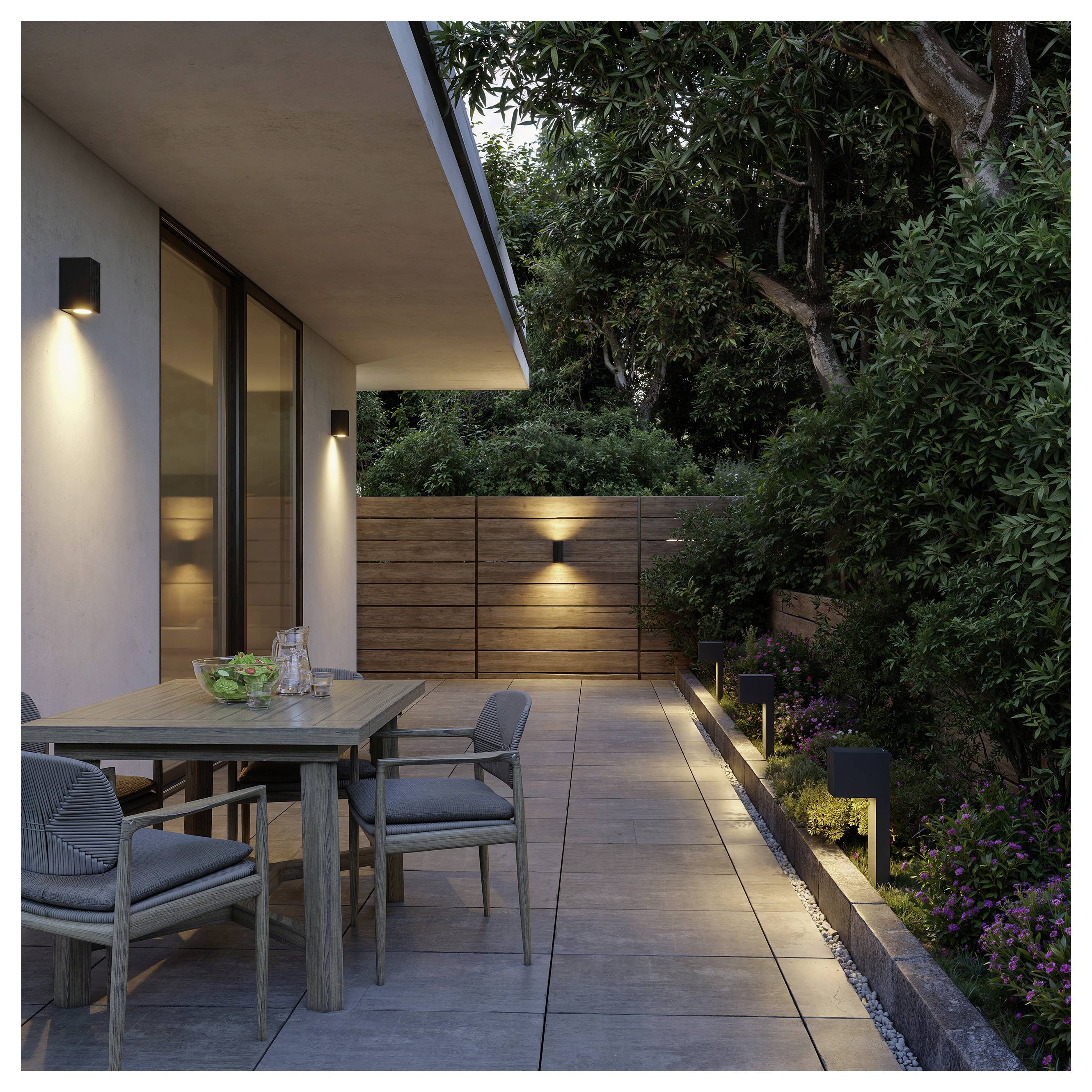 Modern outdoor patio with dining table and chairs beside a house, lit by wall and path lights, bordered by wood fencing and lush trees.<br>