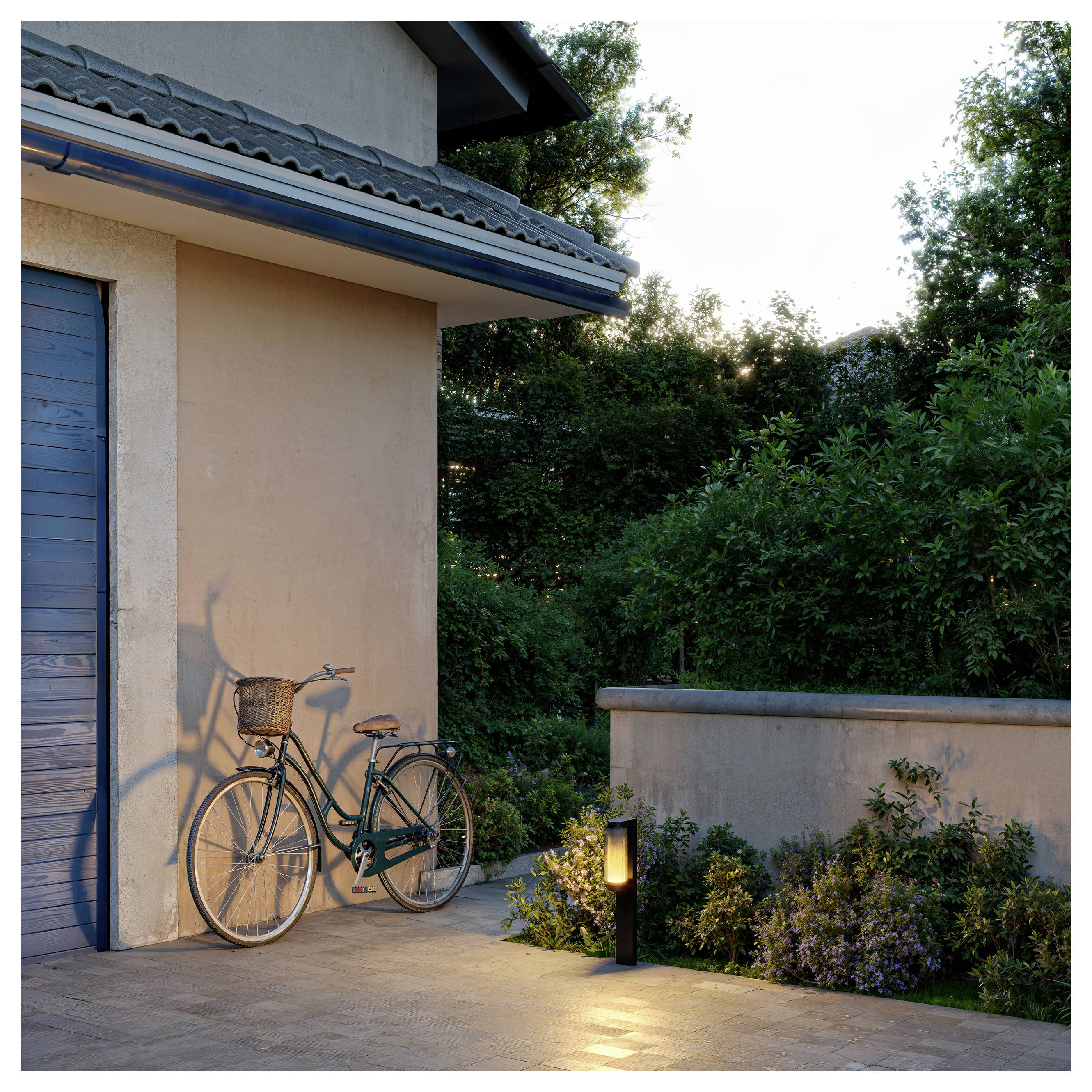 Vintage bicycle leaning against a house wall on a patio, with a lit garden path lamp, low plants, and trees at dusk.