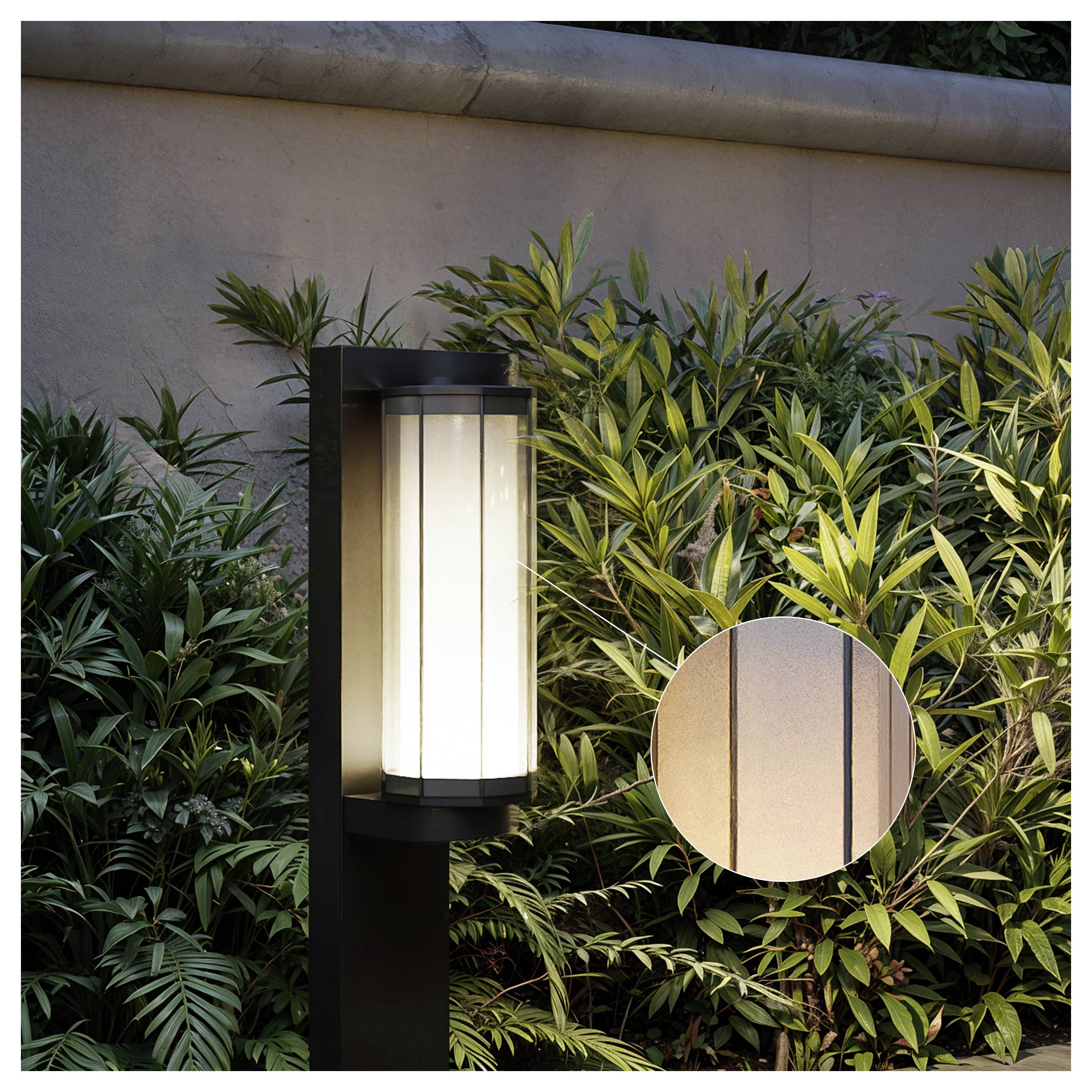 Black outdoor post light with glowing frosted glass beside dense green shrubs and a beige wall; circular inset shows close-up detail of the light fixture.<br>