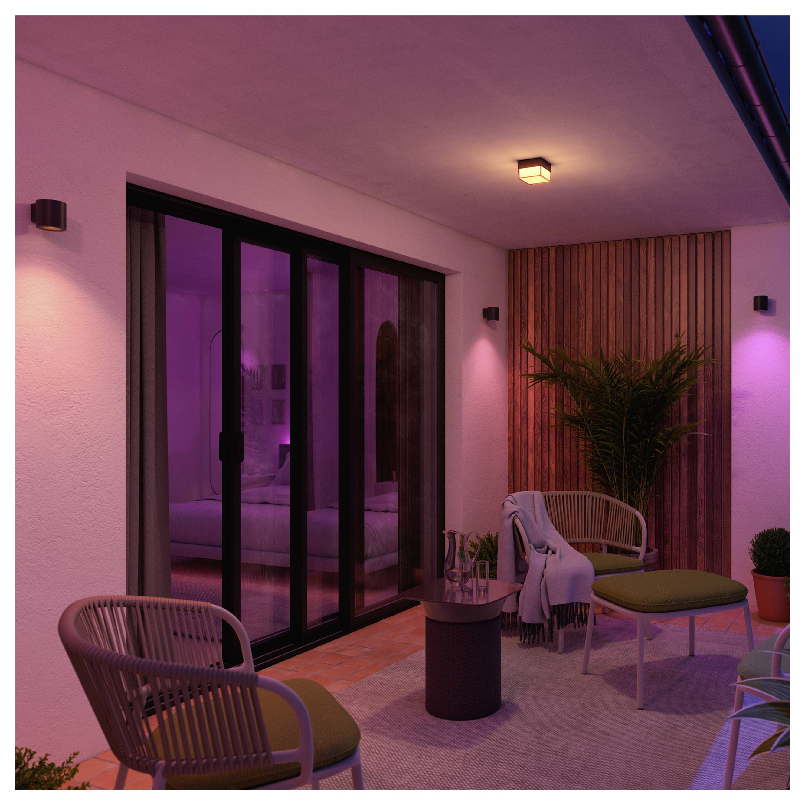 Modern covered patio with lounge chairs, small table, and plants, lit by pink-purple ambient lighting; sliding glass doors open to an interior room.