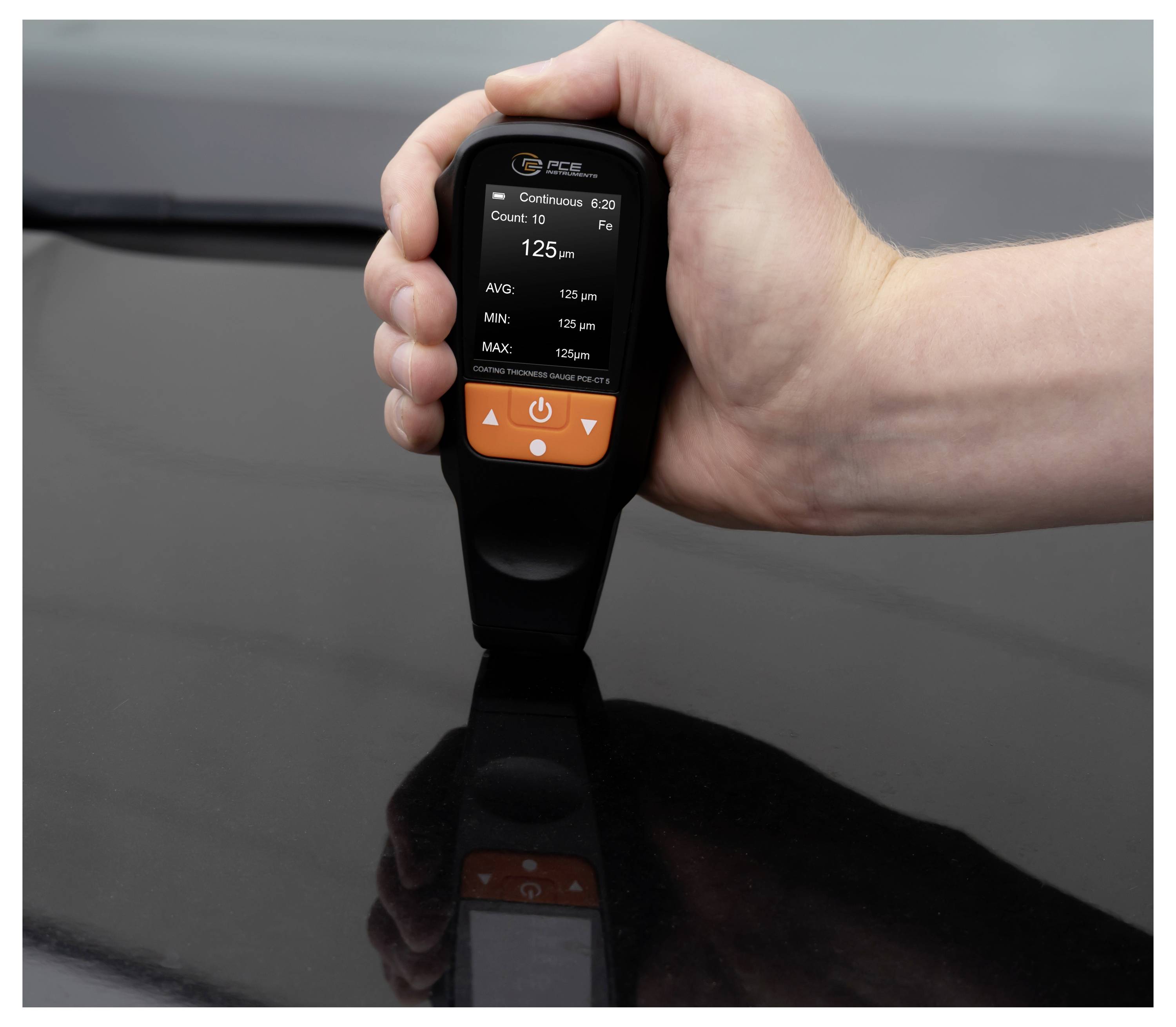 Hand holding a digital paint thickness gauge on a car hood, showing a 125 µm reading on its display.<br>