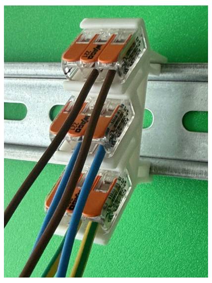 Three wire connectors mounted on a metal rail, joining brown, blue, and yellow-green electrical wires on a green background.