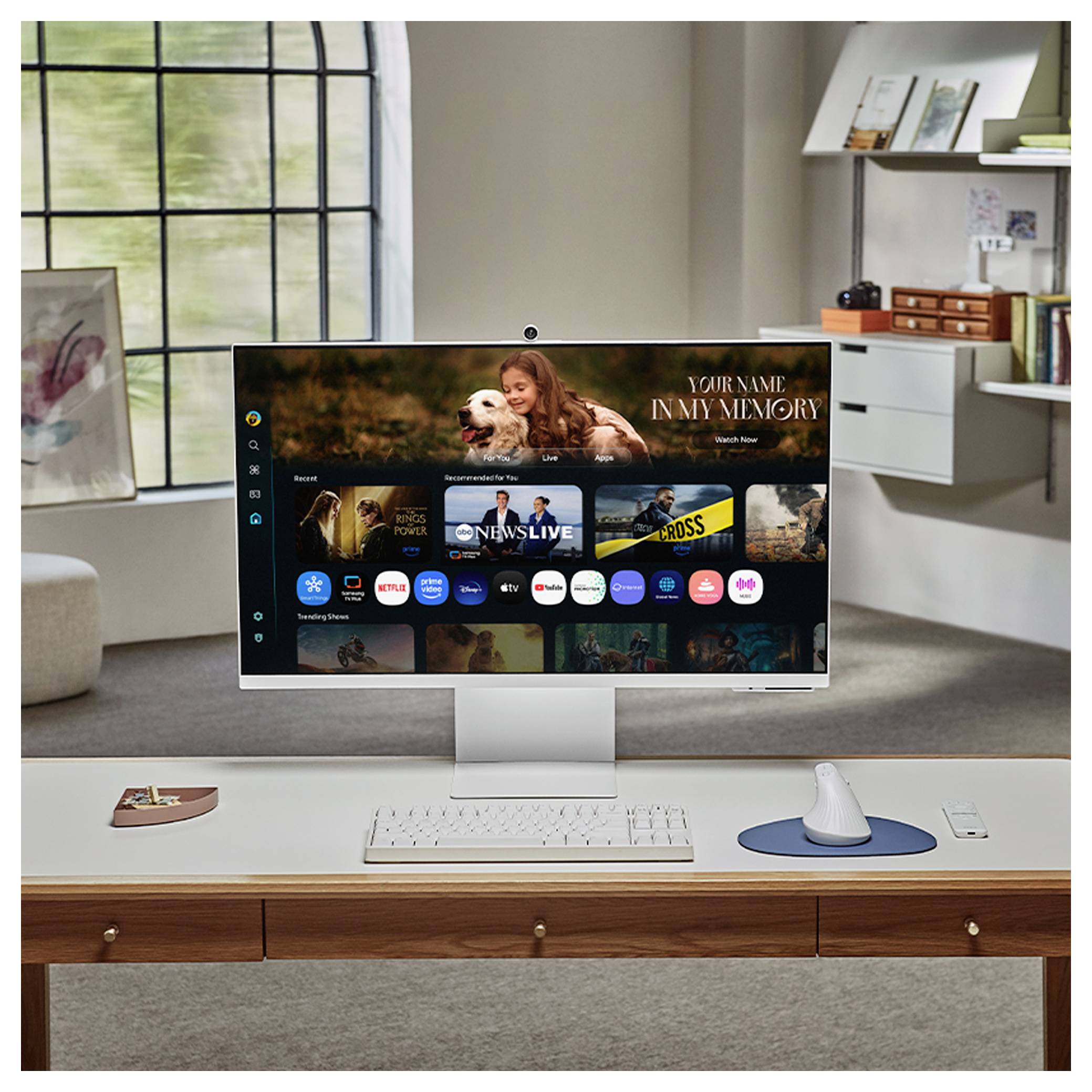 White desktop monitor on a home office desk displays a streaming TV interface with thumbnails and a 'YOUR NAME IN MY MEMORY' banner; keyboard and mouse are below.<br>