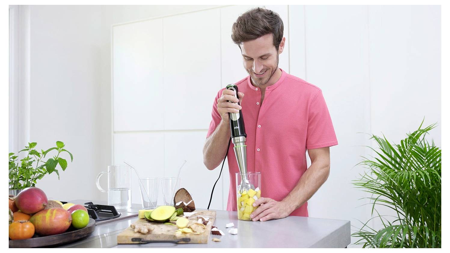 Man using a blender to make a fruit smoothie in a bright kitchen, with cut fruit on the counter and houseplants nearby.