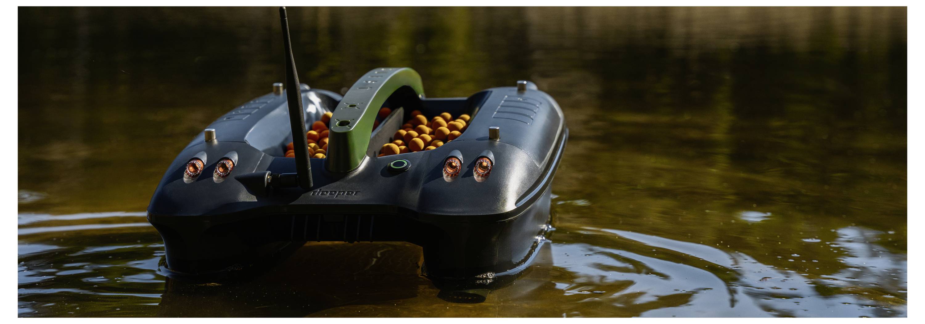 A small black remote-controlled boat with orange bait moves on calm water, likely a fishing bait boat.