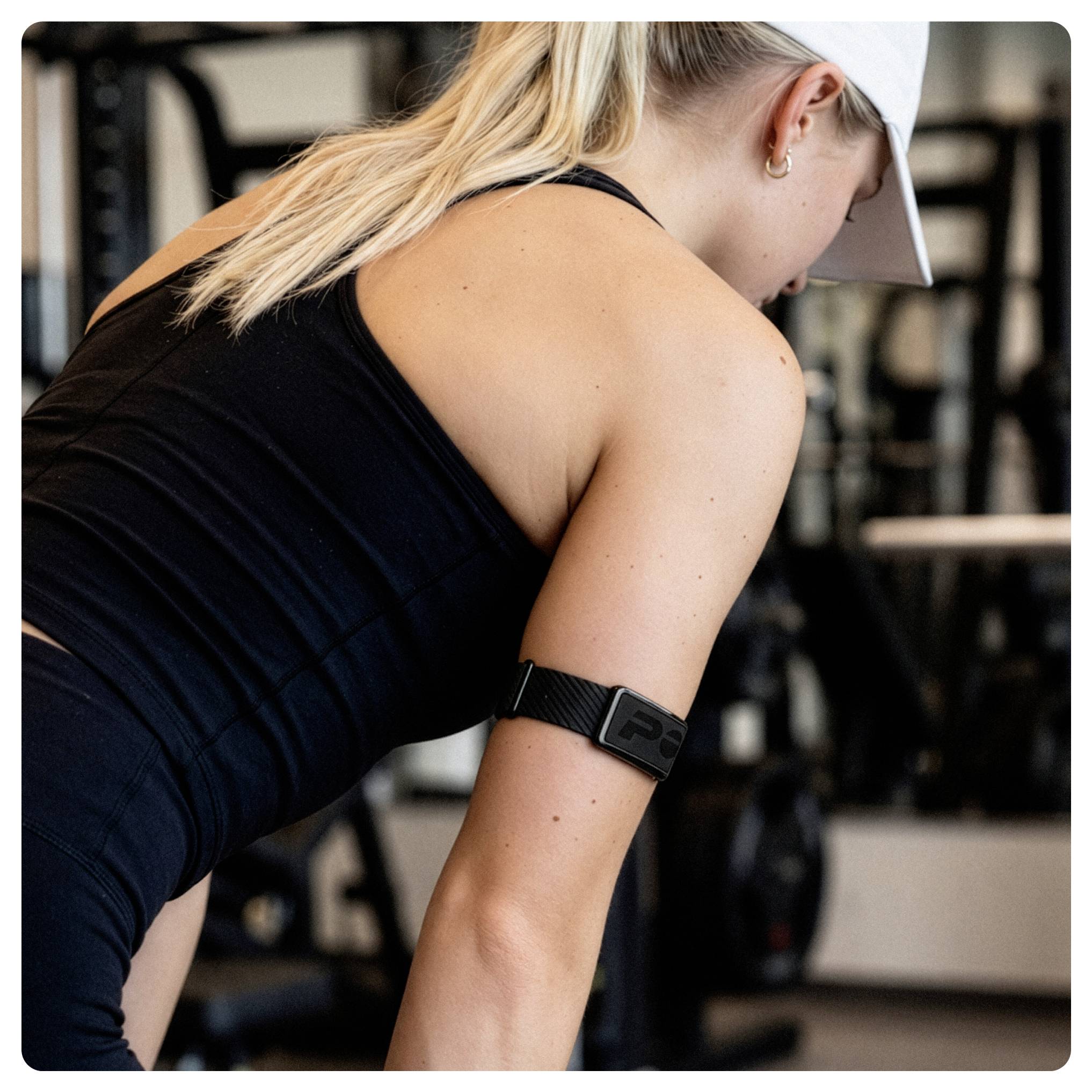 Woman in black athletic wear and white cap leaning forward in a gym, with a fitness tracker on her arm.