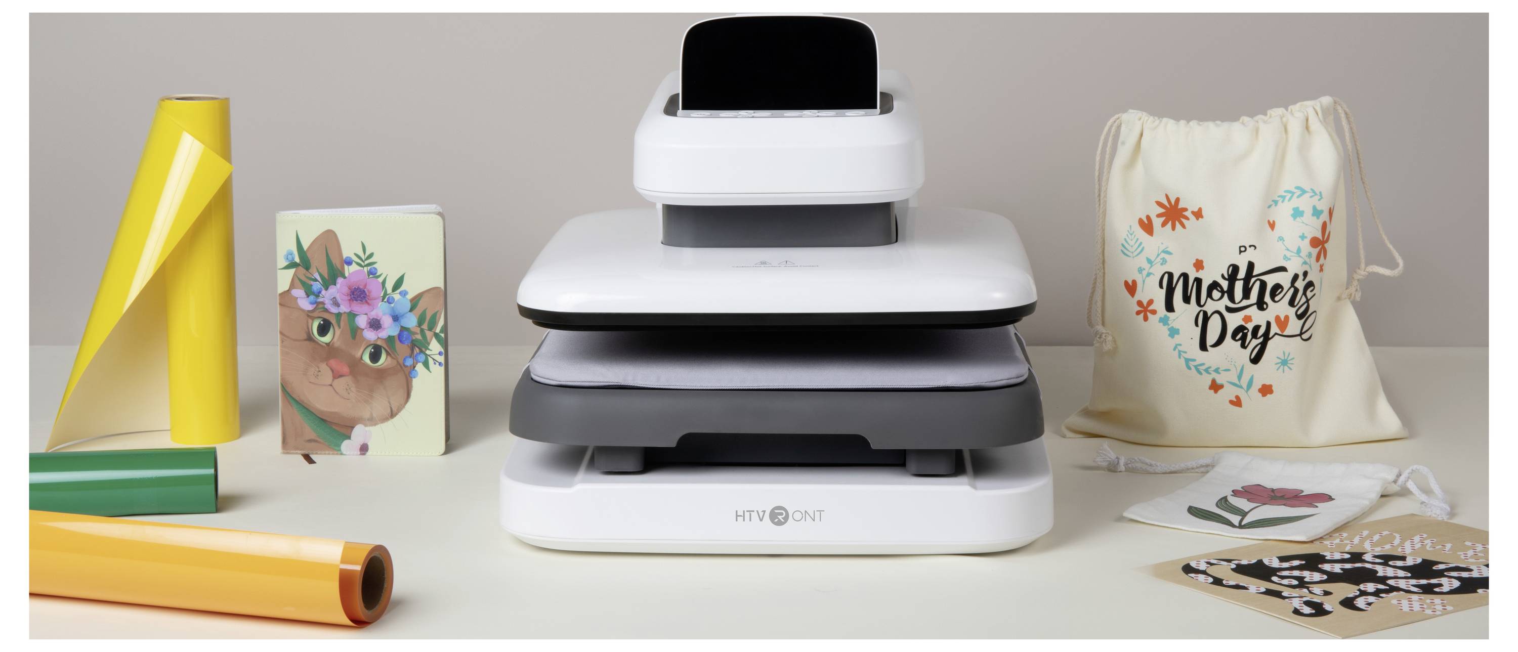 Stack of white HTVRONT heat press machines on a table, surrounded by vinyl rolls, floral transfer sheets, and a 'Mother's Day' gift bag and cards.