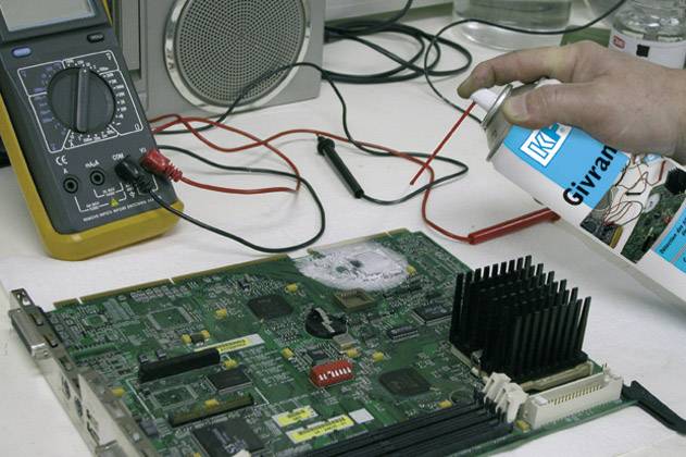 'A person is cleaning a circuit board with compressed air spray. A multimeter and cables can be seen in the background.'