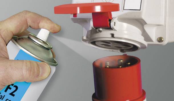 A hand sprays lubricant from an aerosol can onto an electrical plug, with a red cover on top, plug pointing upwards.