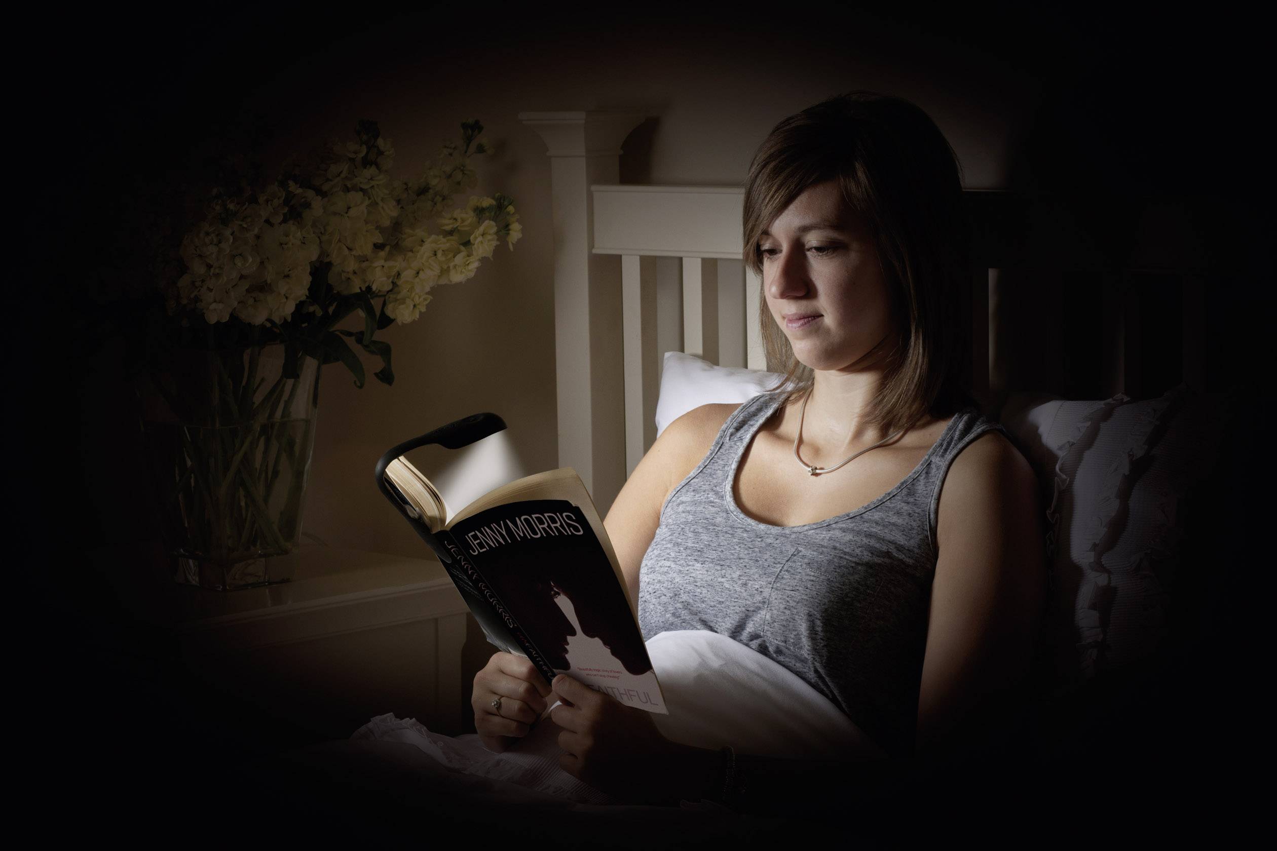 A woman is sitting in bed reading a book. In the background, a vase of flowers stands on a bedside table.