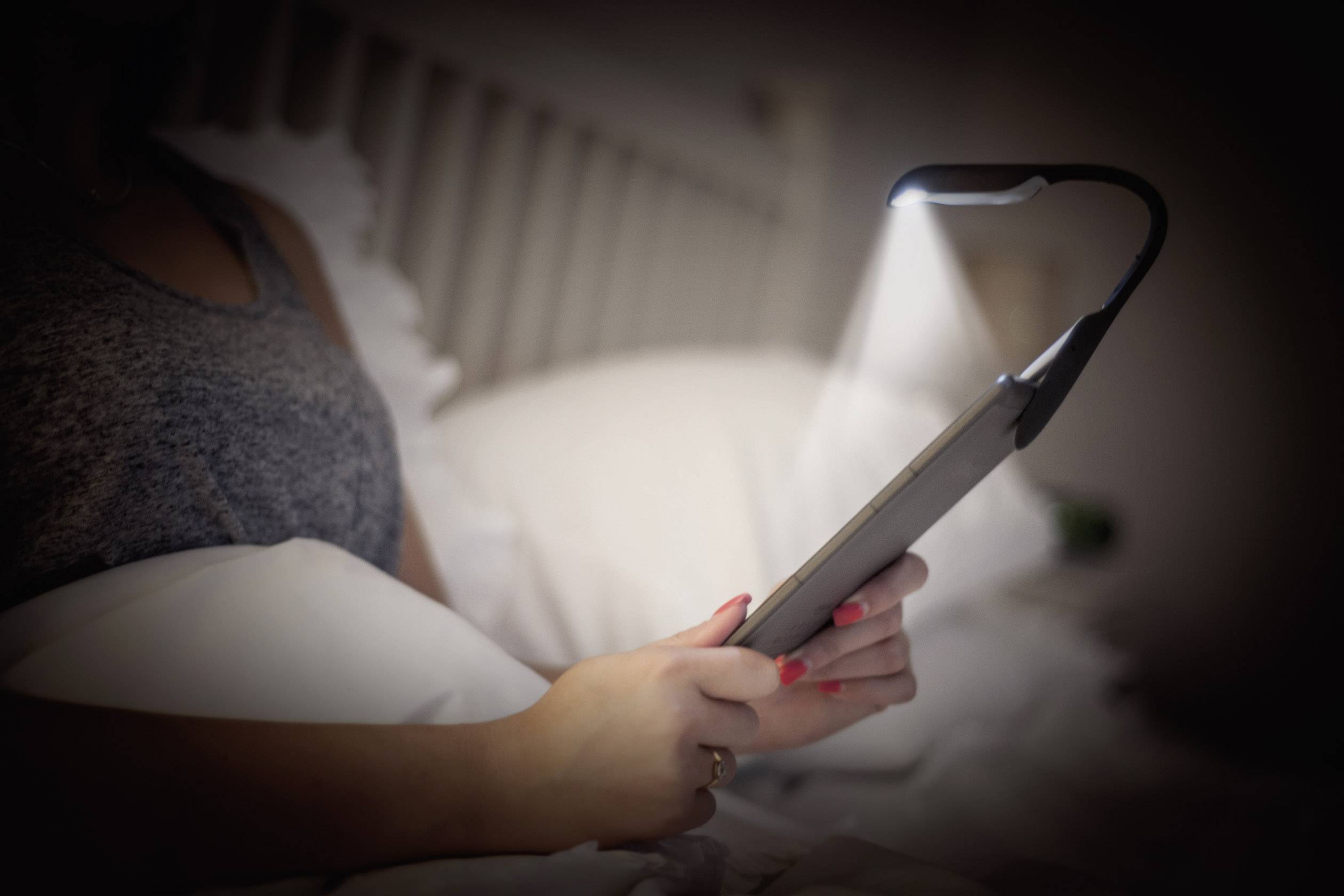 A person is reading on a tablet with an attached reading light while in bed. The surrounding environment is dark, and the light illuminates the screen.