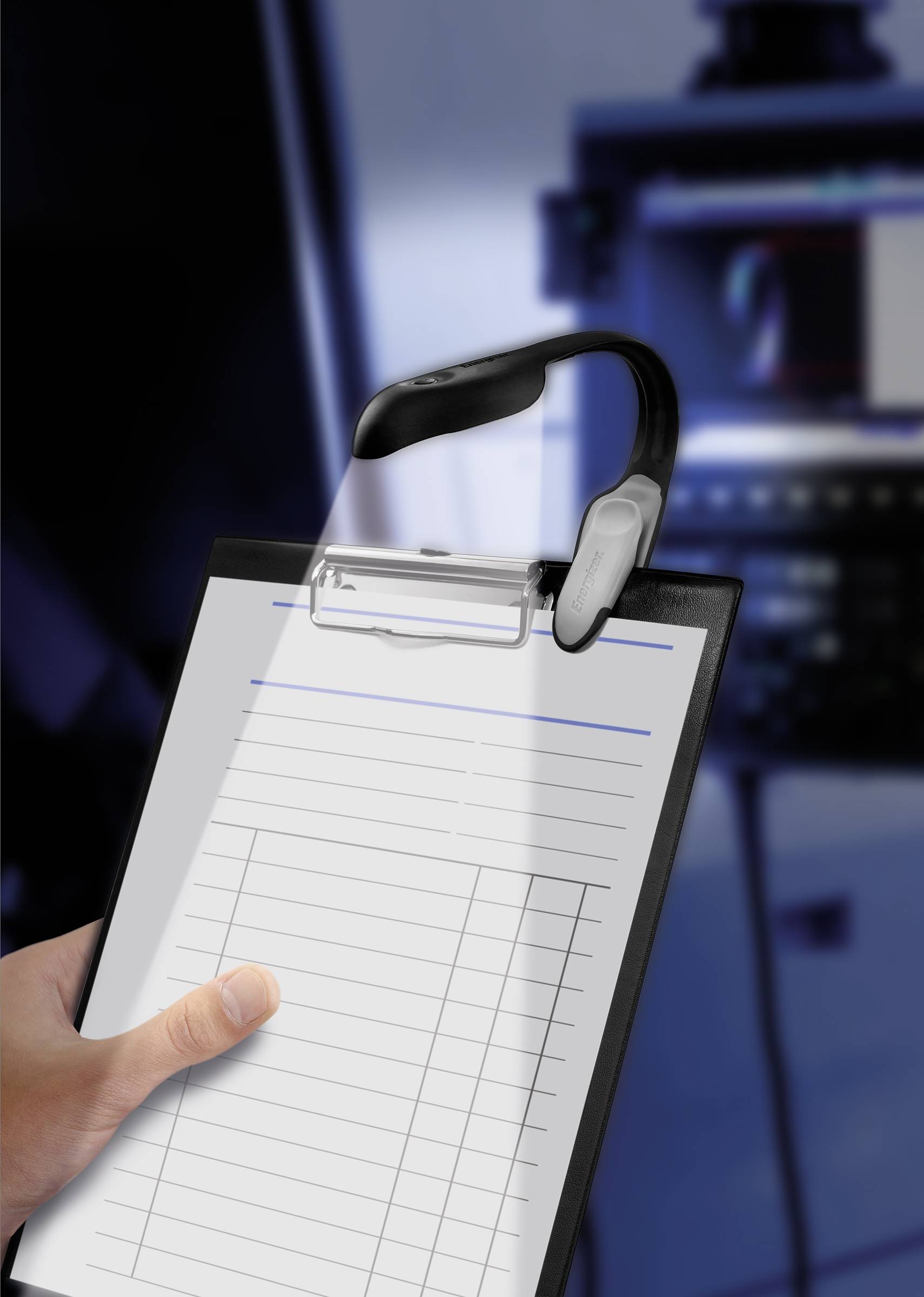 A hand is holding a clipboard with a blank form. A computer screen is blurrily visible in the background.