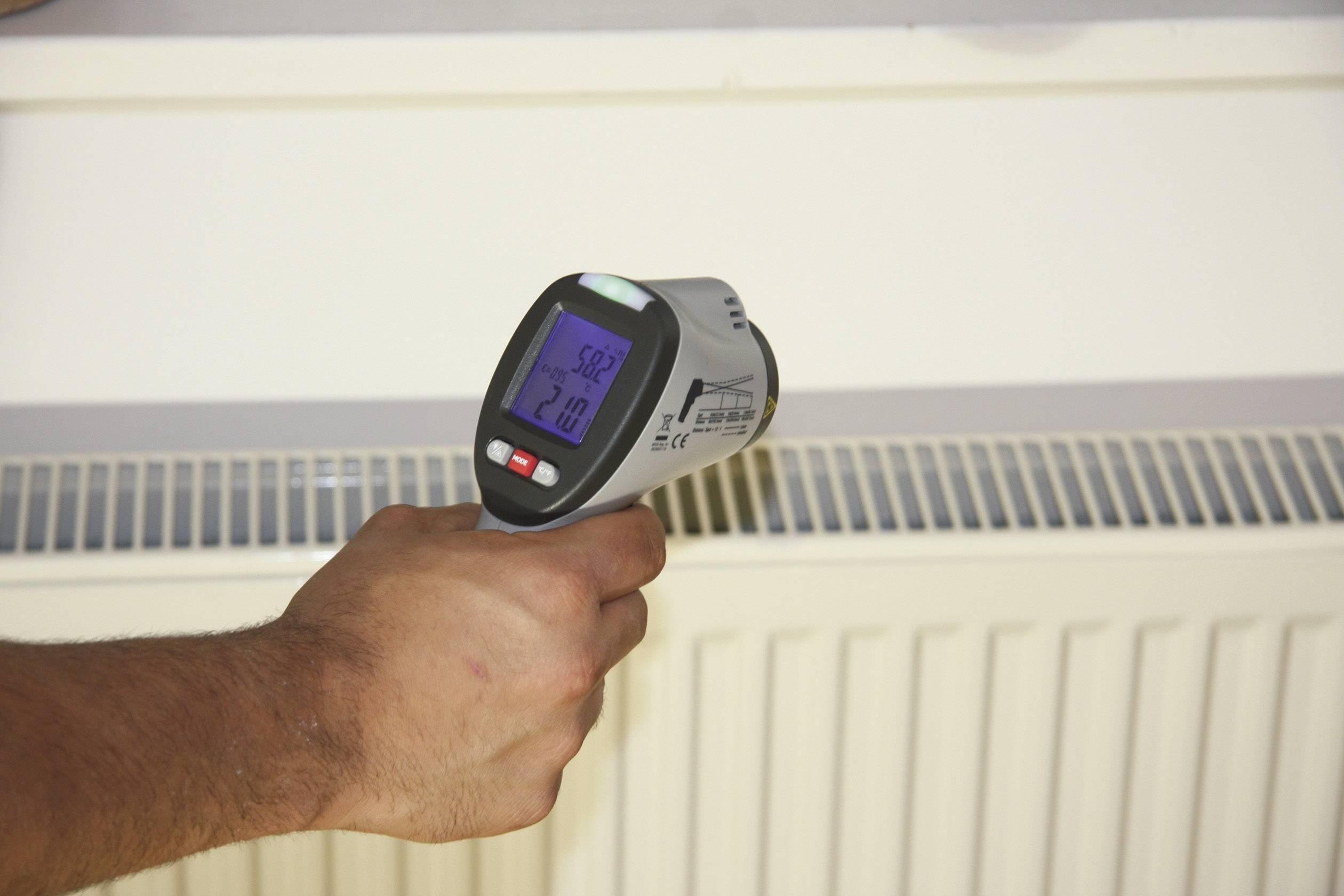 A hand is holding an infrared thermometer measuring the temperature of a radiator. The display shows 52.3 degrees Celsius.