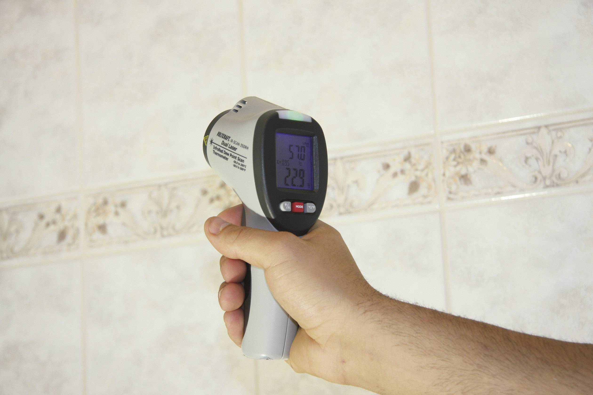 A hand holds an infrared thermometer in front of a tiled wall. The display shows temperature measurements of 5.0 and 22.9 degrees.