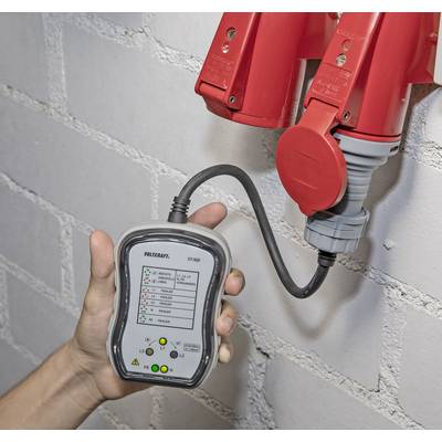 A hand is holding a testing device connected to red industrial sockets to check electrical voltage and phase configuration.