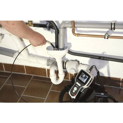 A person is using a drain camera to inspect a drainage pipe. A monitor in the foreground displays the captured image.
