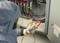 A person in protective clothing is measuring the voltage of an electrical control cabinet using a digital multimeter. Safety precautions are being observed.