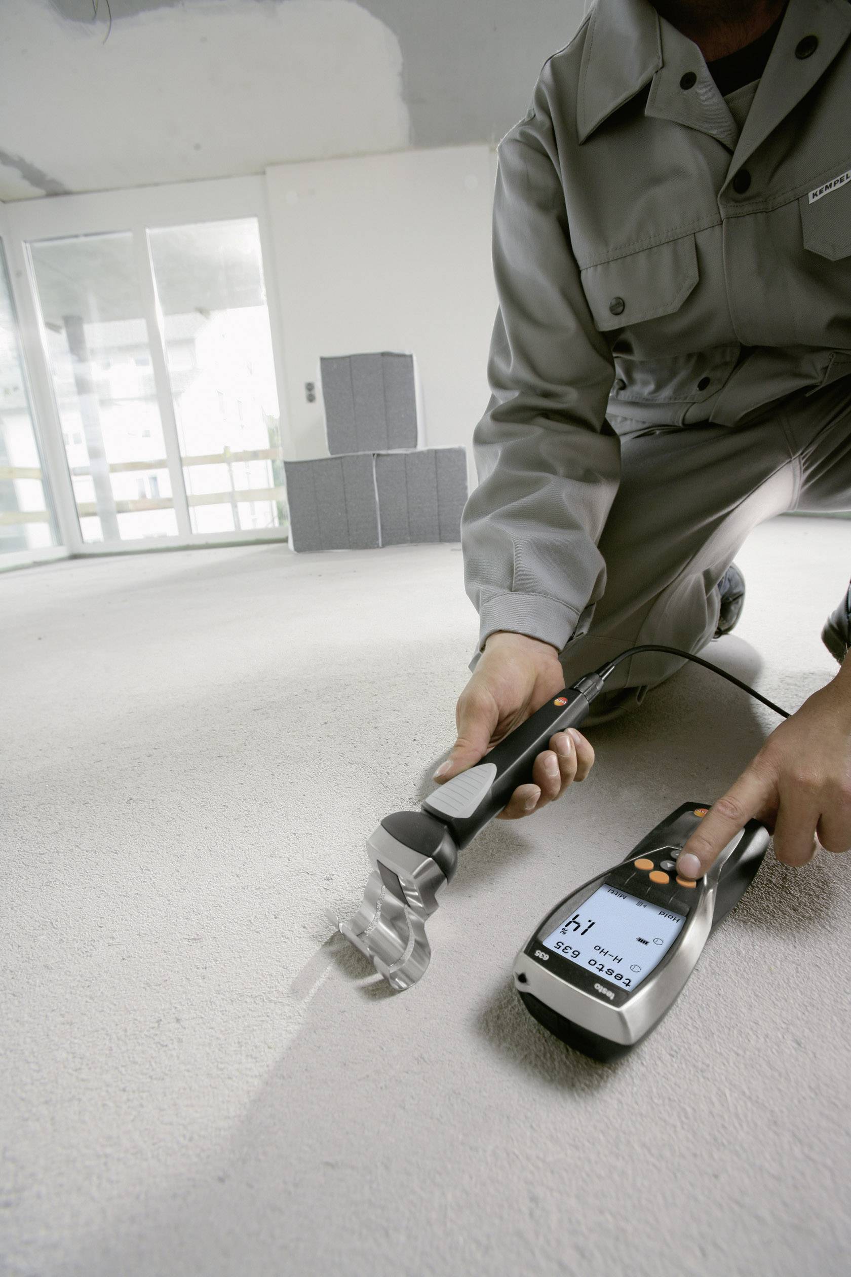 A person is measuring the moisture of a concrete floor with an electronic device in an unfinished room. Cardboard boxes are standing in the background.