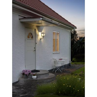 A white house facade at dusk with an illuminated front door. A small table and two chairs are placed in front of it, surrounded by flowers.