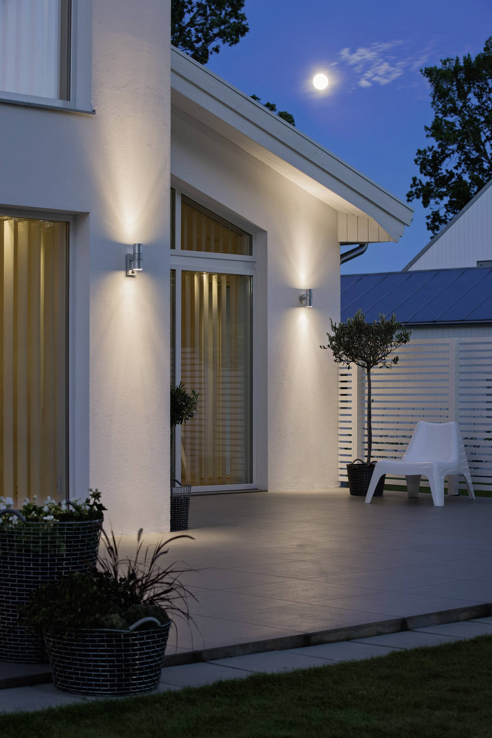 External view of a modern house at night with wall lights switched on, a terrace, and a full moon in the background.