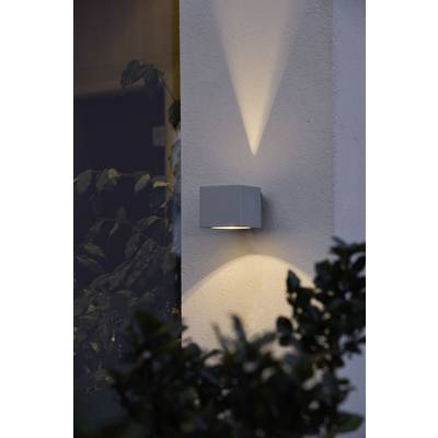 An exterior wall light that casts warm light onto the wall. Illuminates part of the façade and an adjacent bush.