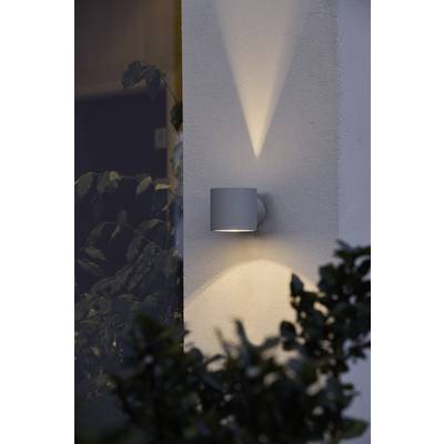 Wall light at a building corner, casting soft illumination upwards and downwards. Plants are visible in the foreground.