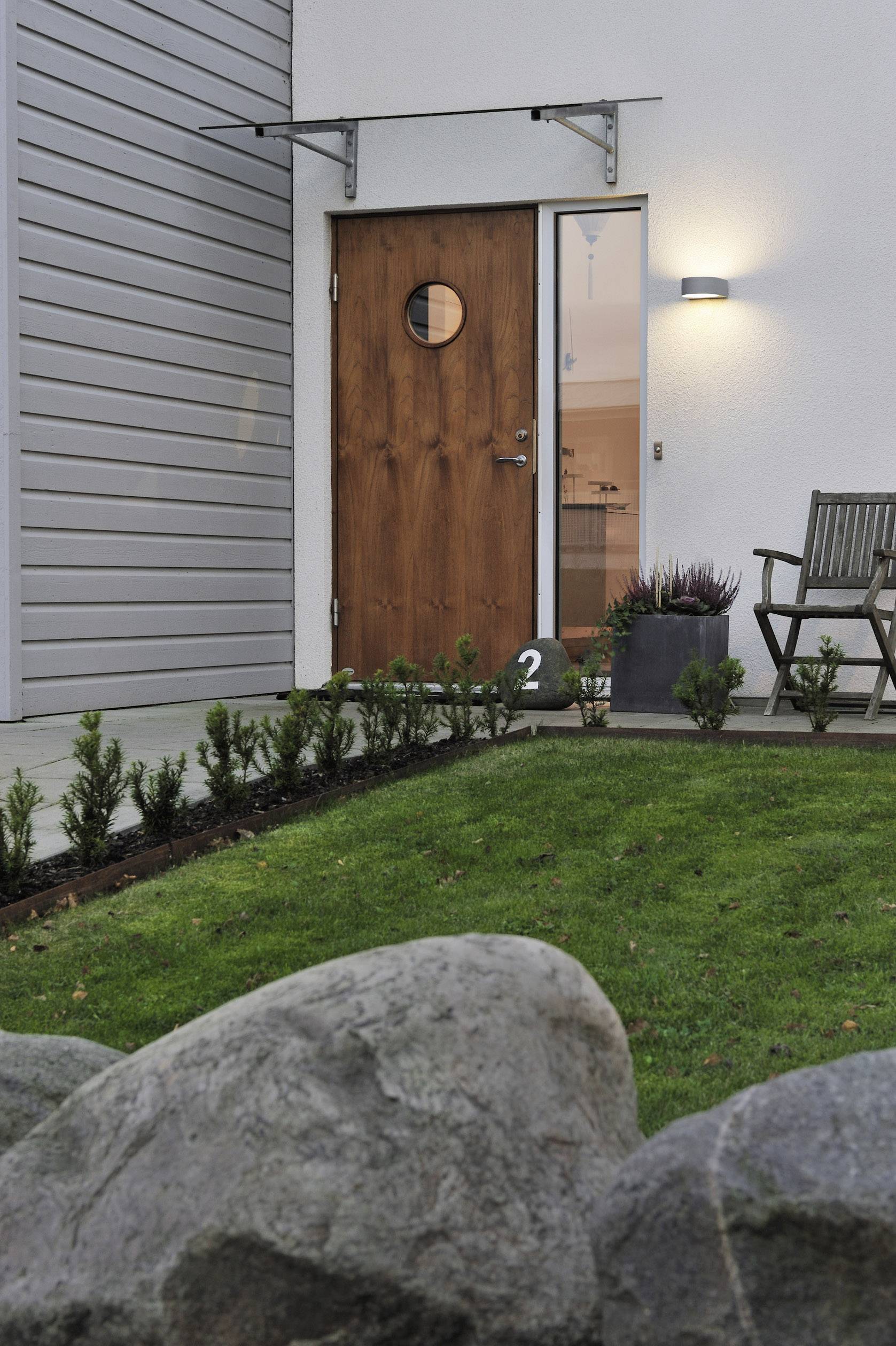 Wooden door on a modern house facade with a circular window and garden in the foreground. Narrow cladding on the left, stone path with chairs on the right.