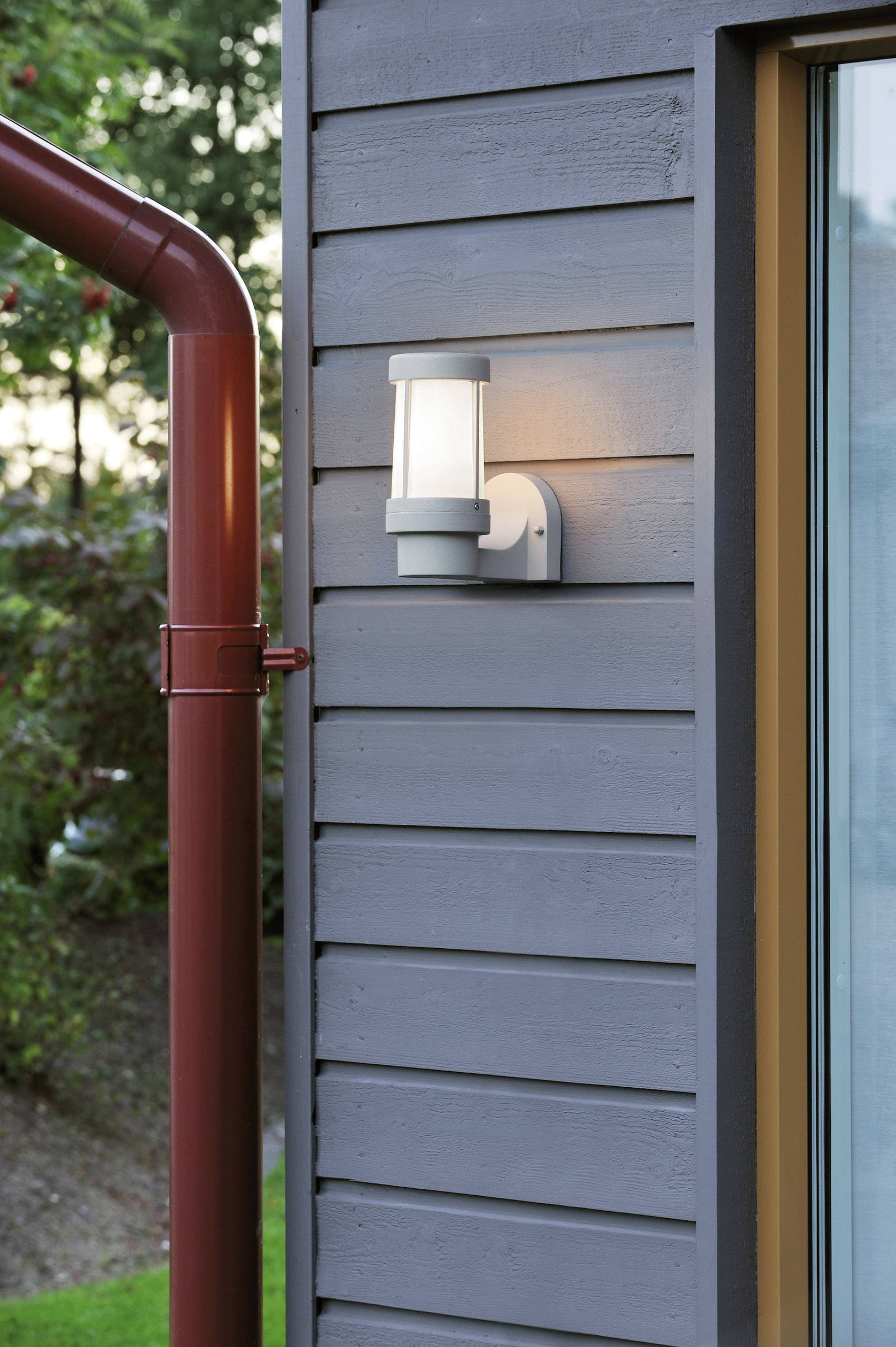 Exterior view of a modern grey house wall with a glowing wall light and a red downspout.
