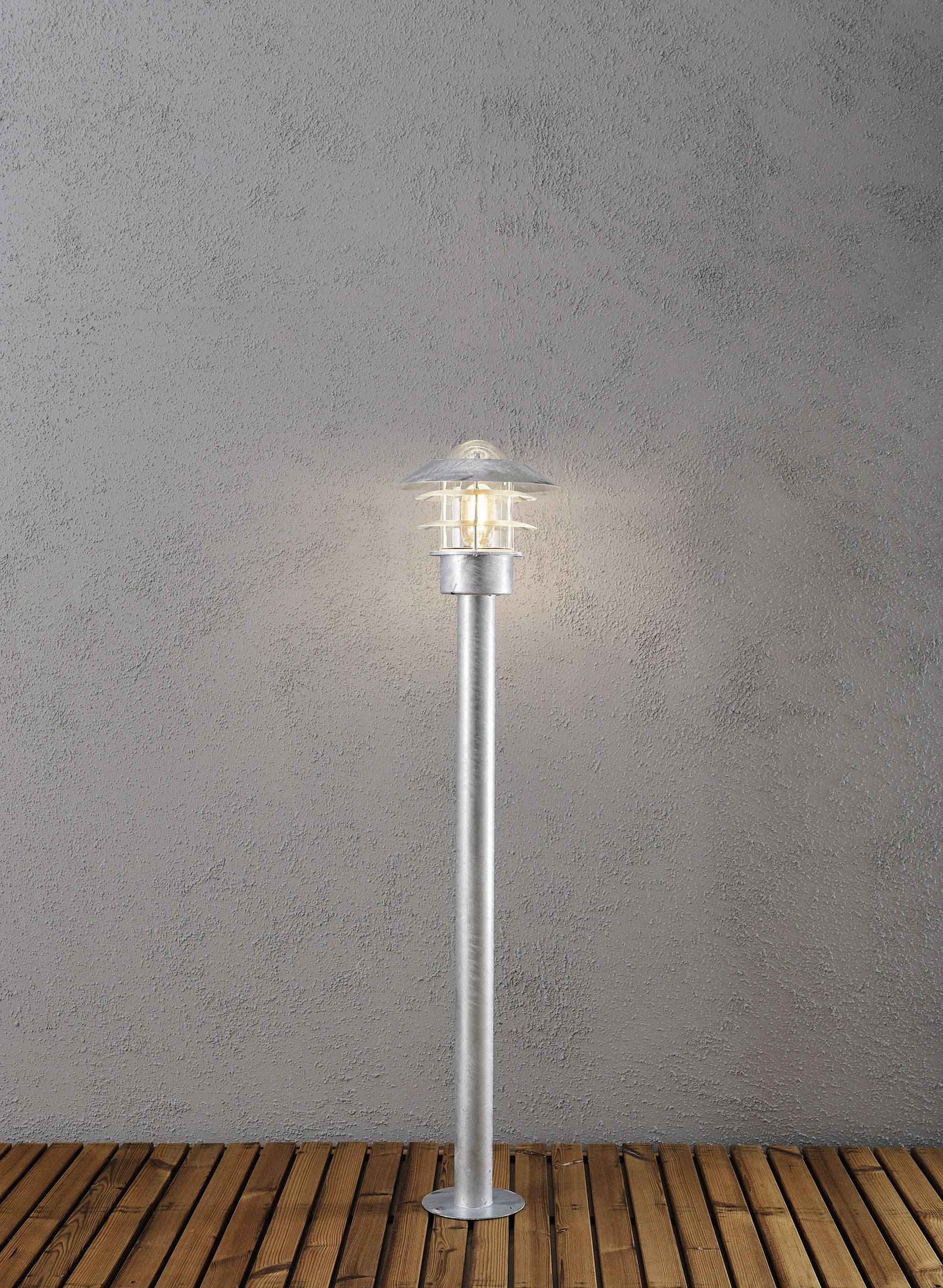 A modern street lamp stands on a wooden floor in front of a grey wall. The lamp is illuminated and emits a soft light.