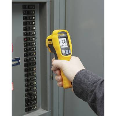 A hand holds a yellow infrared thermometer in front of an electrical control cabinet, displaying 32.2 degrees Celsius on the screen.