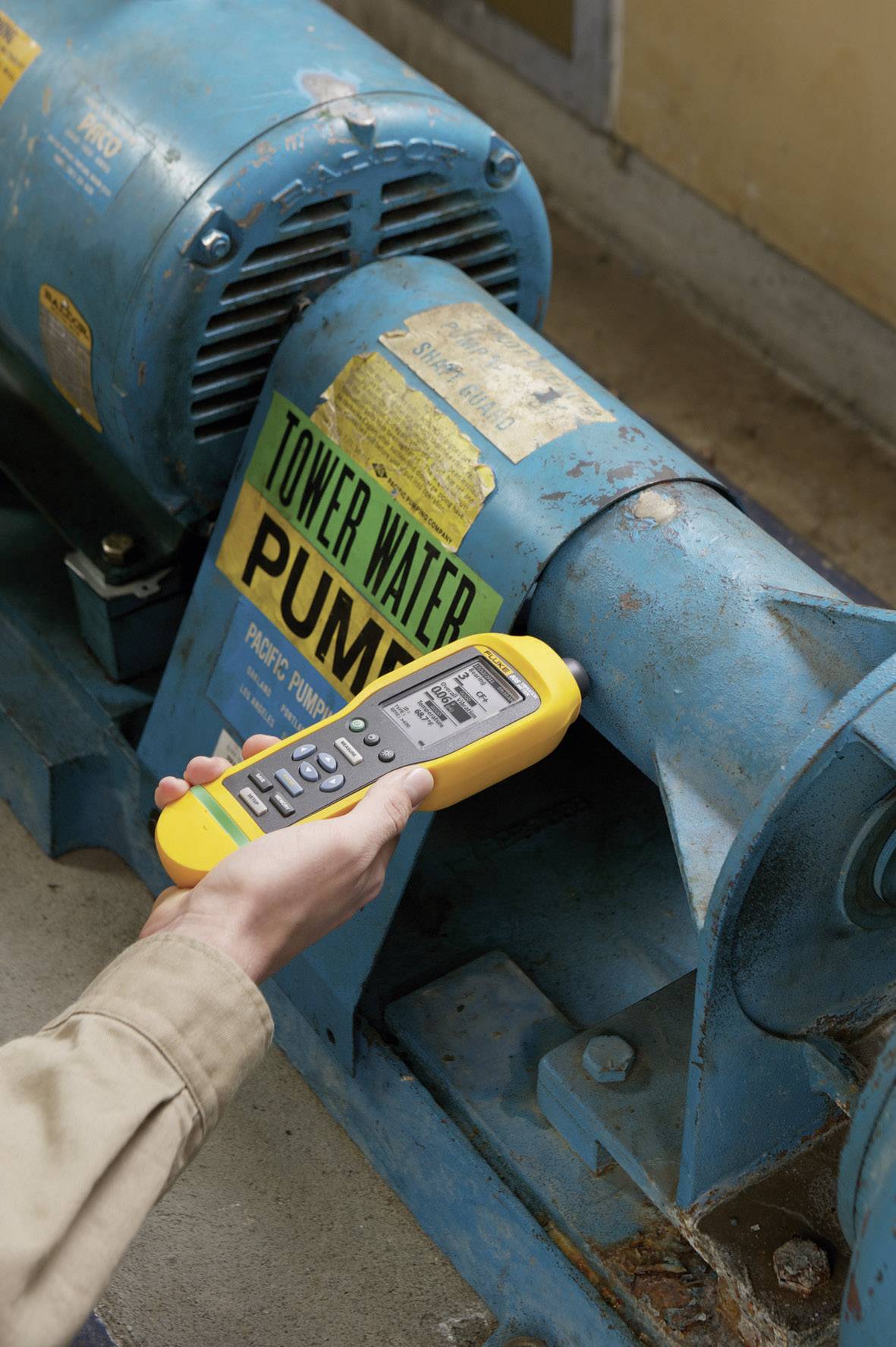 A hand is holding a yellow measuring device, checking the vibration of a blue industrial machine with a green 'PUMP' sign attached.