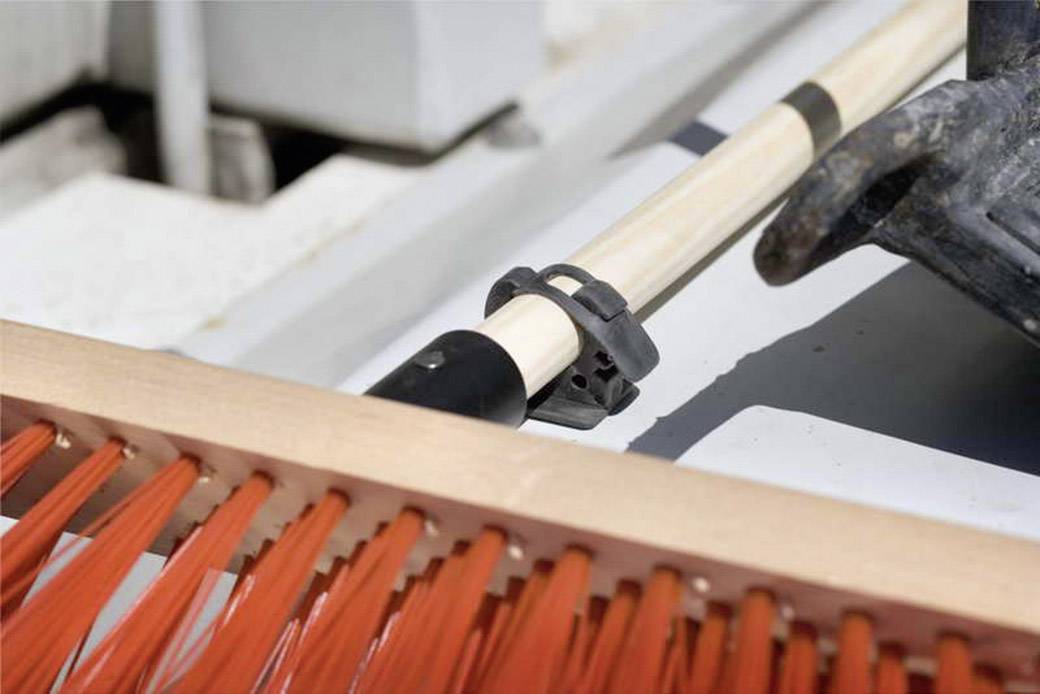 A broom with orange bristles rests on a wooden handle, secured with a mounting bracket, against a blurred background.