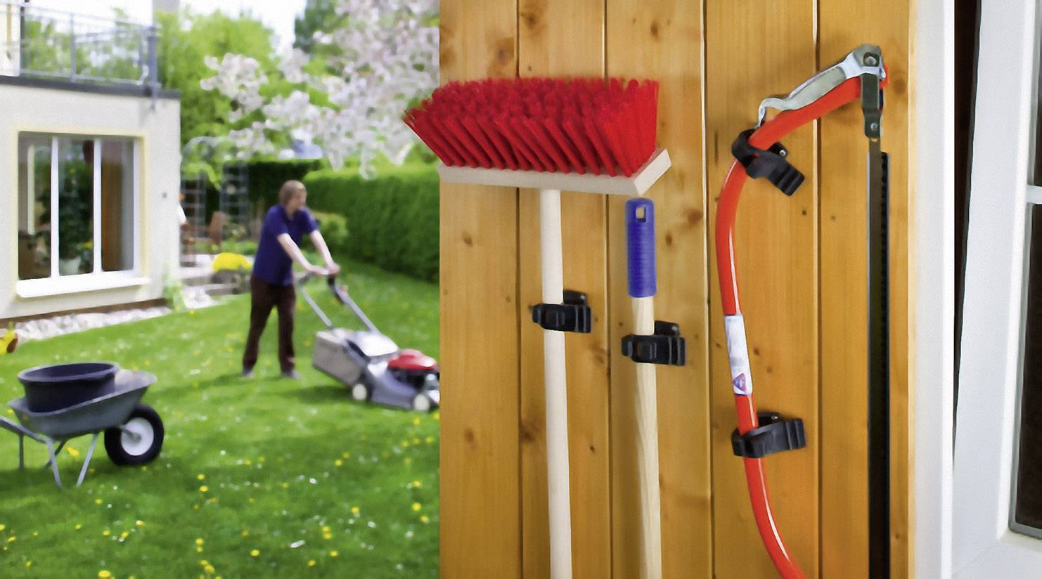 A person is mowing the lawn in the garden. In the foreground, garden tools such as a broom and a saw are hanging on a wooden lath.