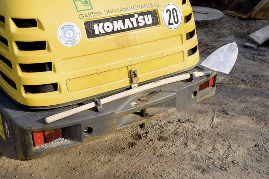 A small yellow excavator with 'Komatsu' and 'Landscaping' written on it, with a garden and landscaping background. A shovel is resting on the back of the vehicle.