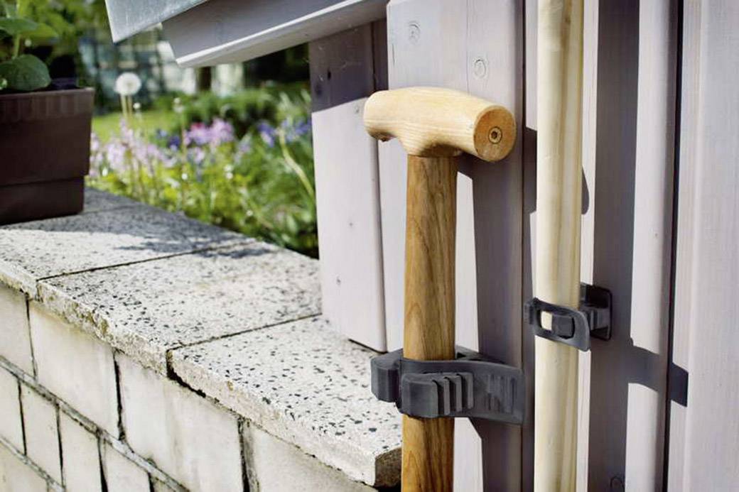 A walking stick and a broom are hanging on a house wall, with a garden and flower bed in the background.