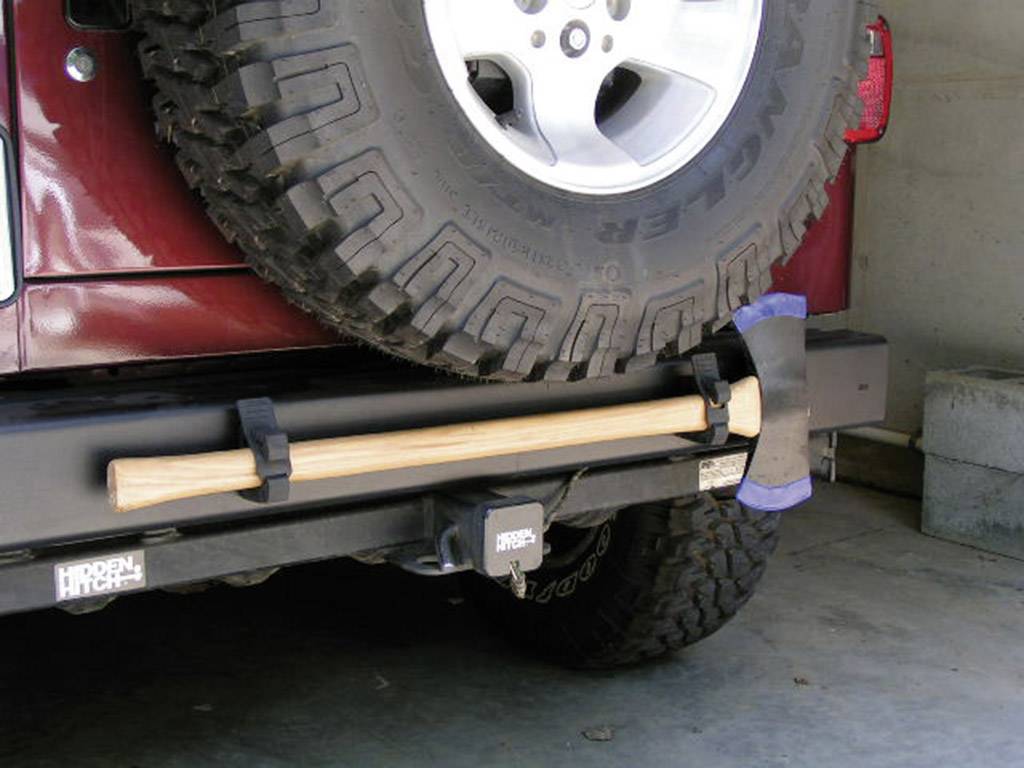 A spare tyre is mounted on the rear of a car. Underneath, an axe with a wooden handle is secured to a holder.