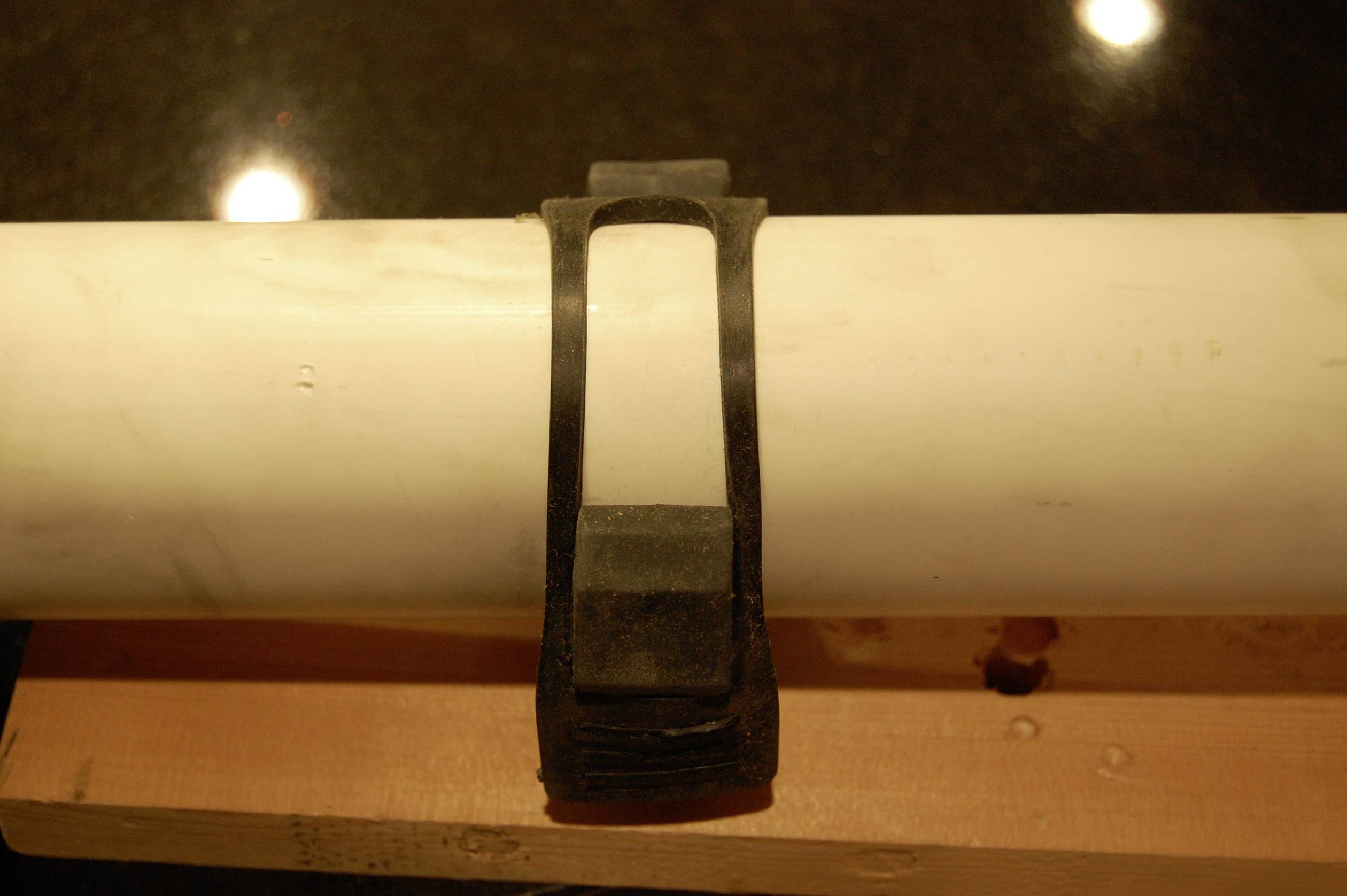 A black plastic clip fastens a large white pipe to a wooden block, which is illuminated from above.