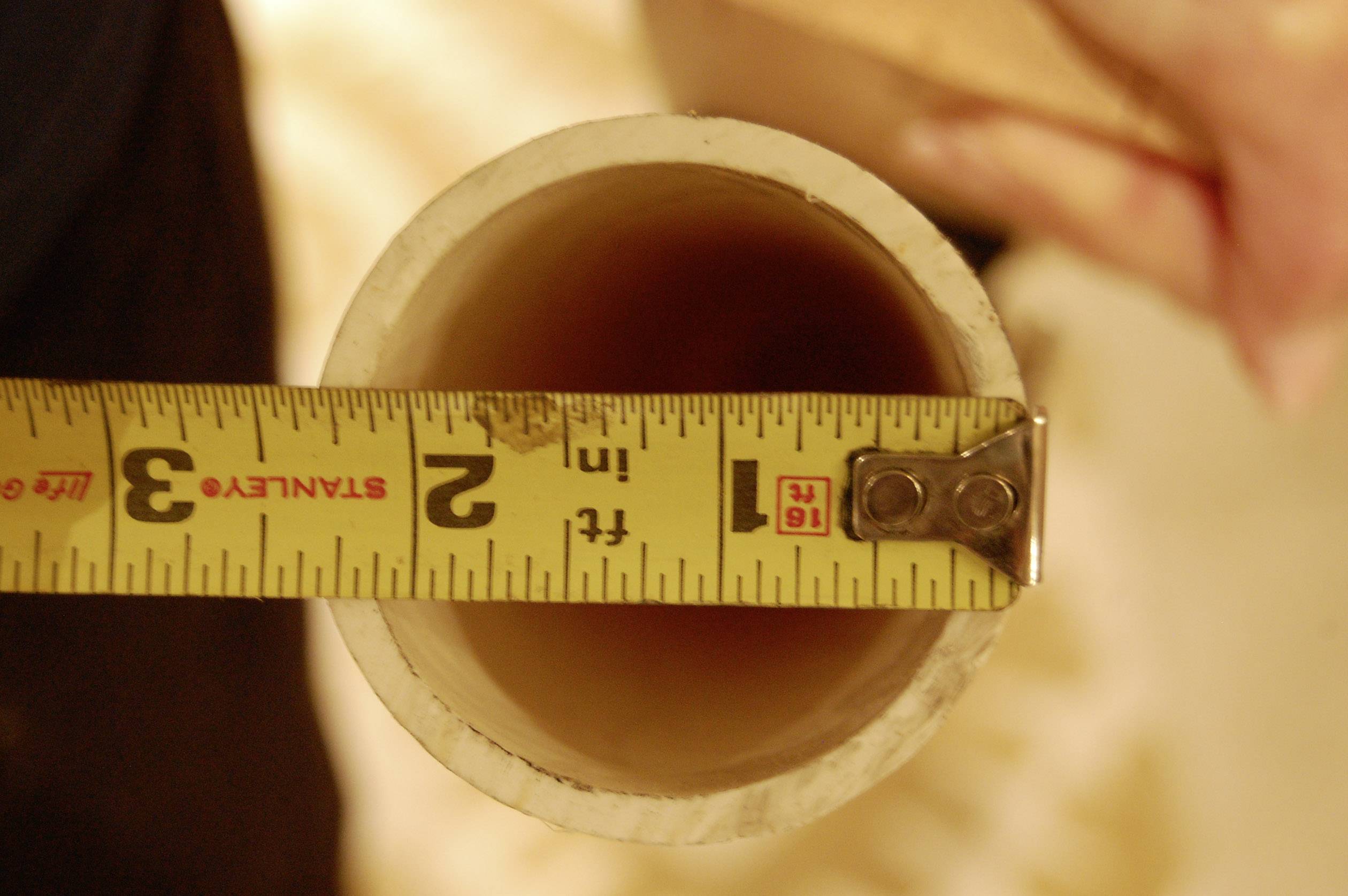 A person is measuring the diameter of a plastic pipe with a metal tape measure, which indicates approximately 7.6 cm.
